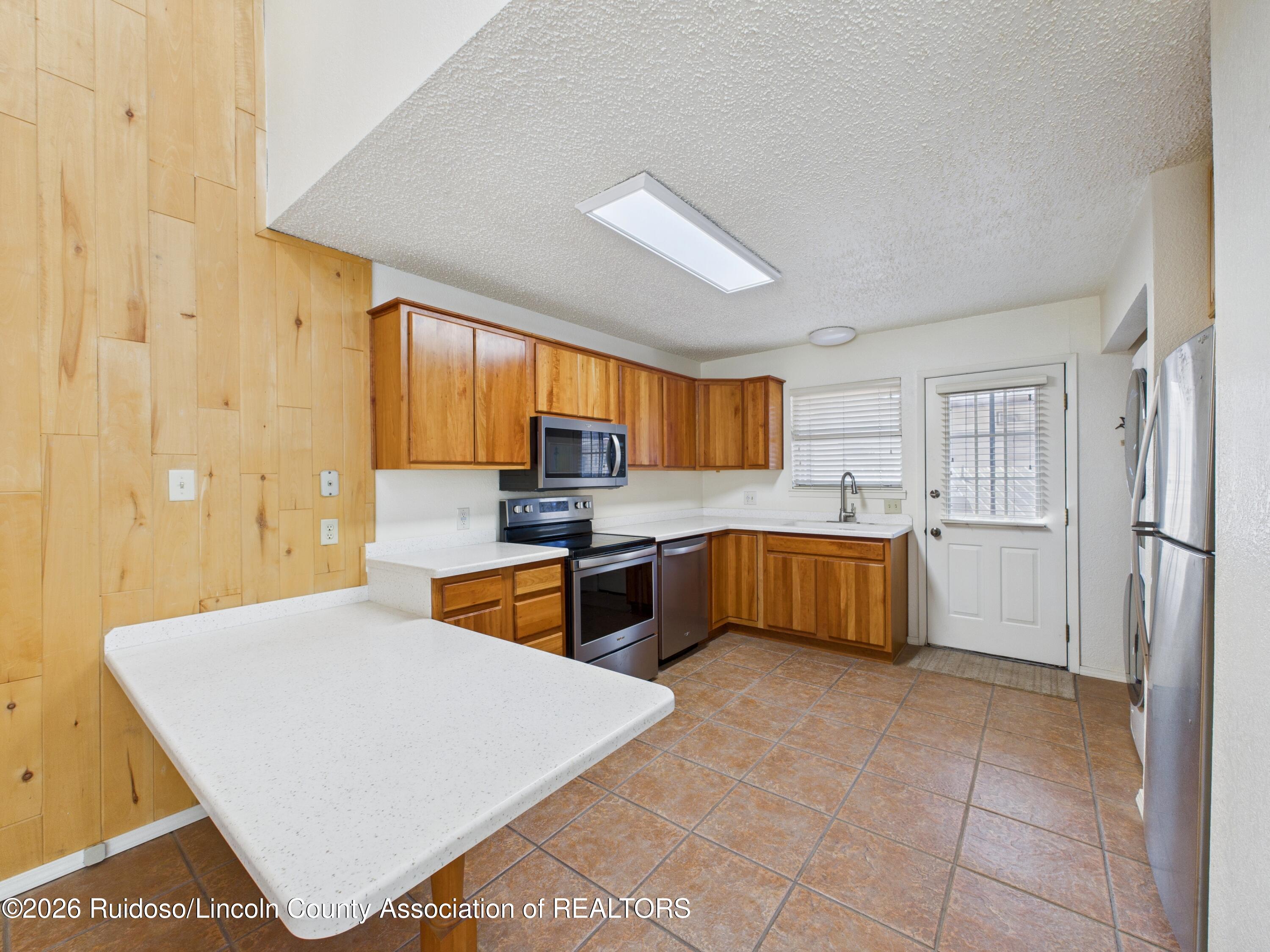 Kitchen with wood cabinetry, white peninsula countertop, stainless steel appliances, and terracotta tile floor.