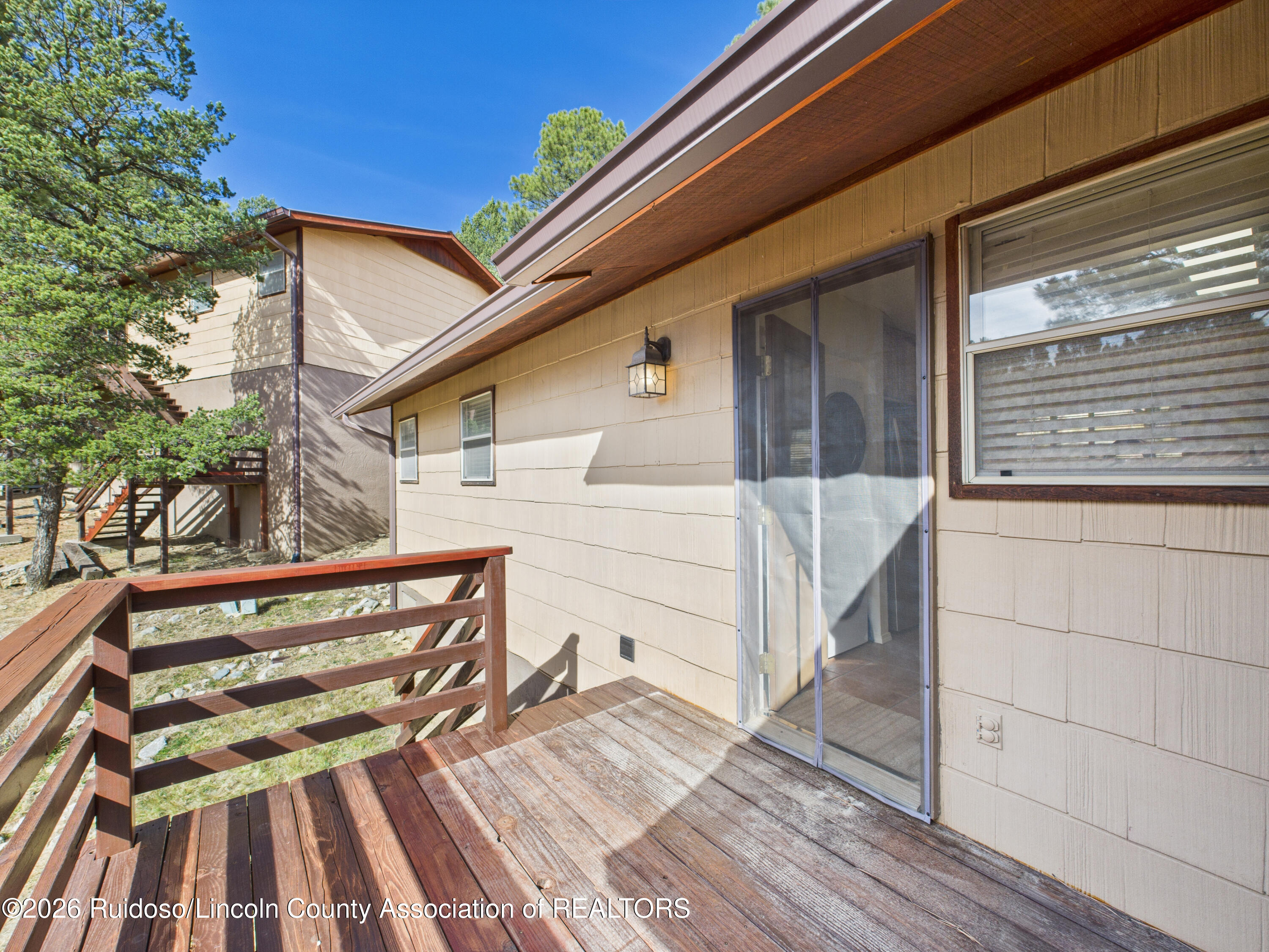 Wooden deck beside a beige home, with a sliding screen door and pine trees nearby.