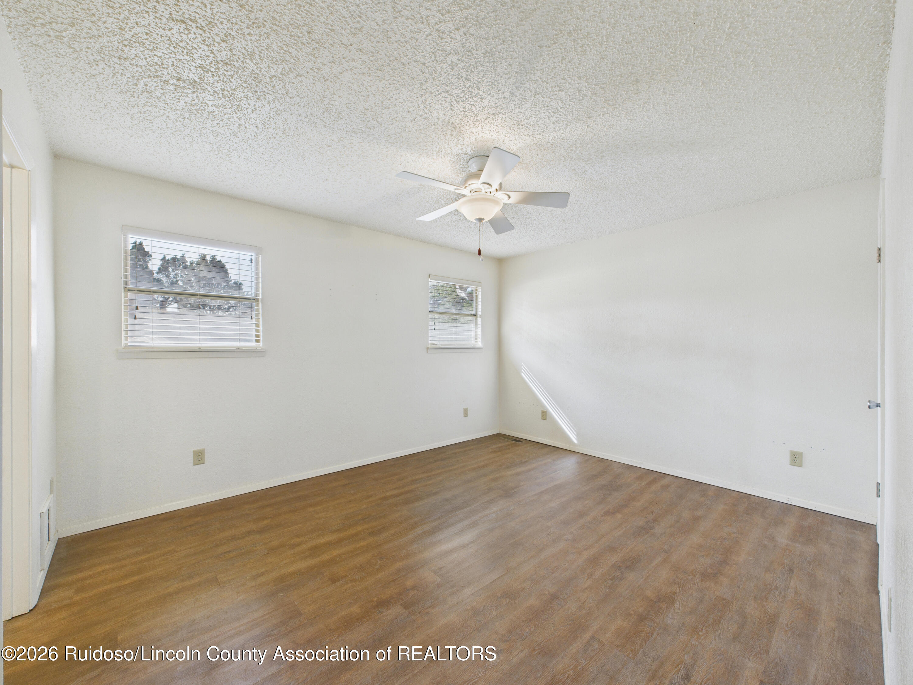 Empty room with brown wood-look flooring, two windows with blinds, and a white ceiling fan.