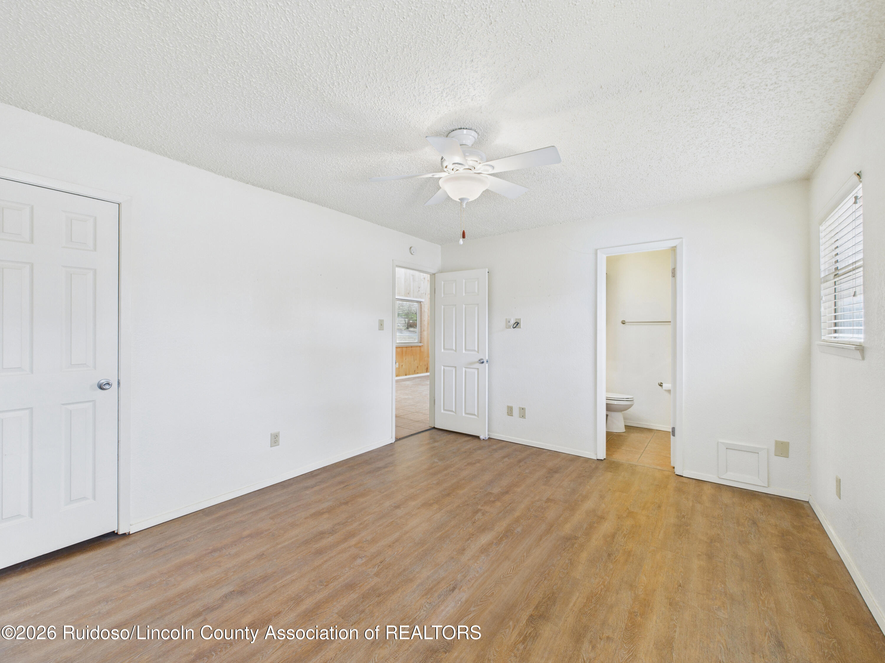 Empty bedroom with wood-look floors, ceiling fan, and open bathroom doorway.