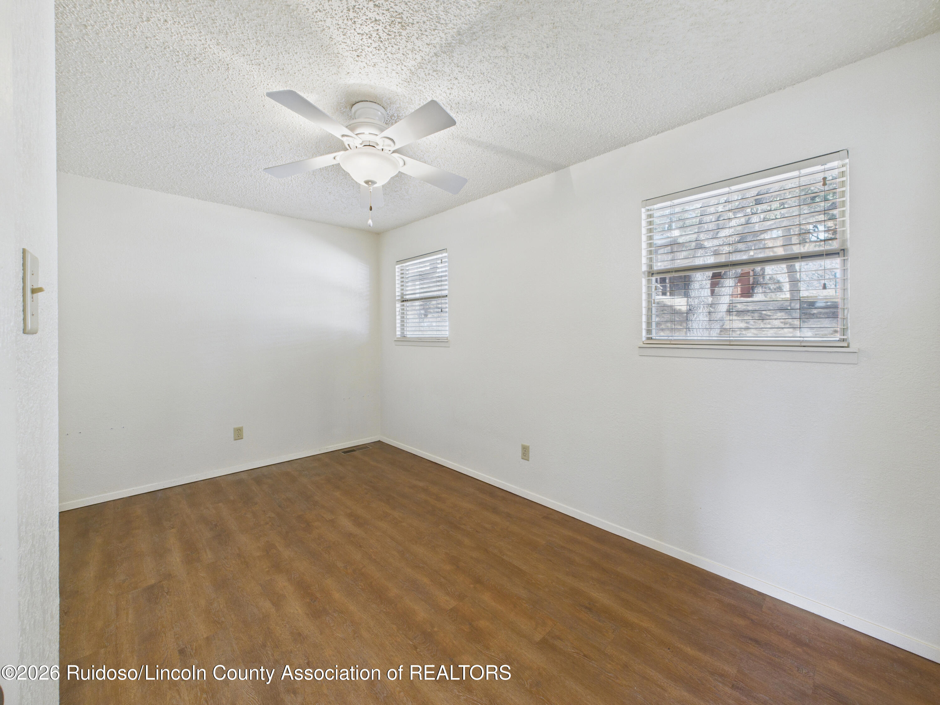 Empty room with brown flooring, two windows with blinds, and a ceiling fan.