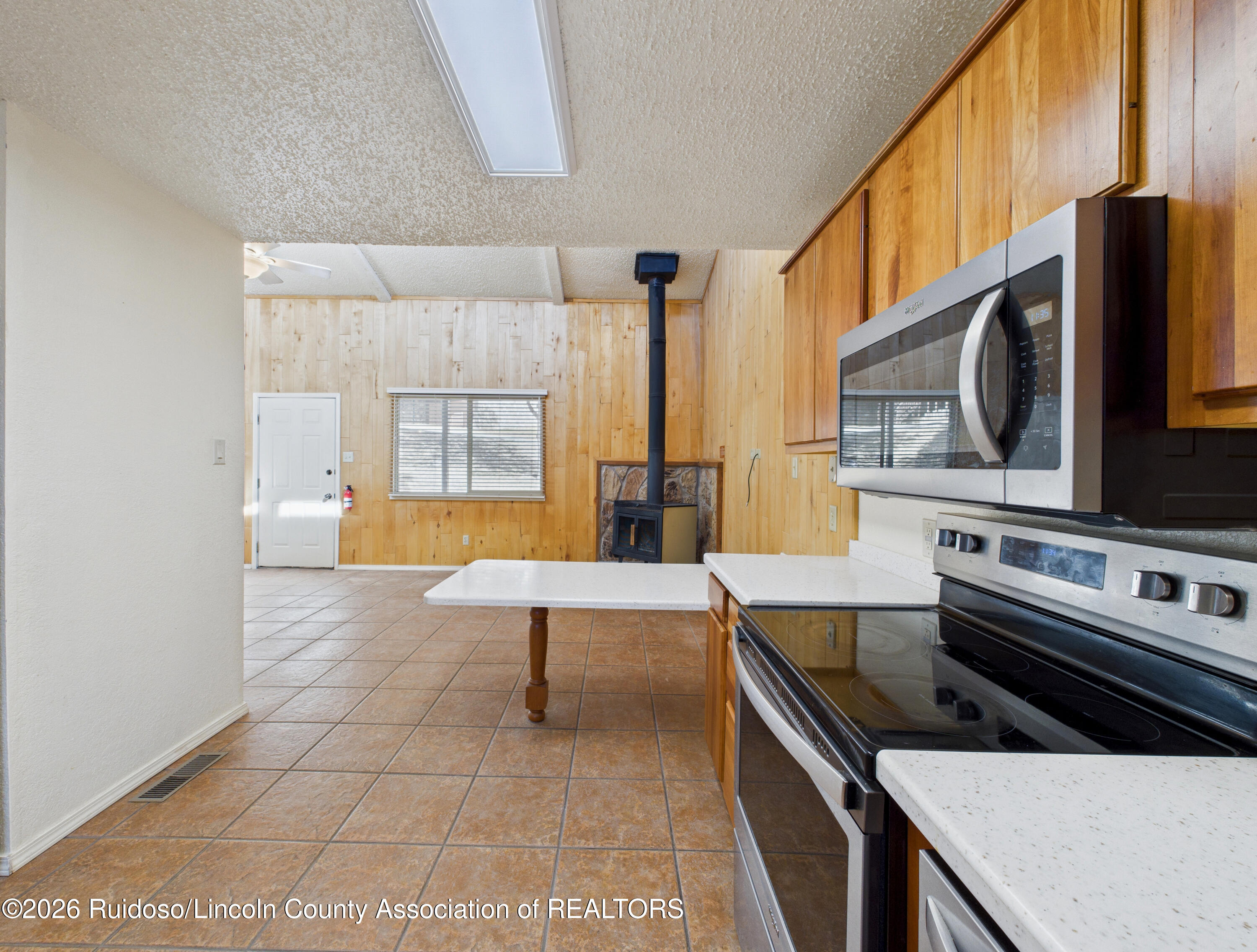 Kitchen with wood cabinets and stainless appliances, opening onto a tiled room with a wood-burning stove.