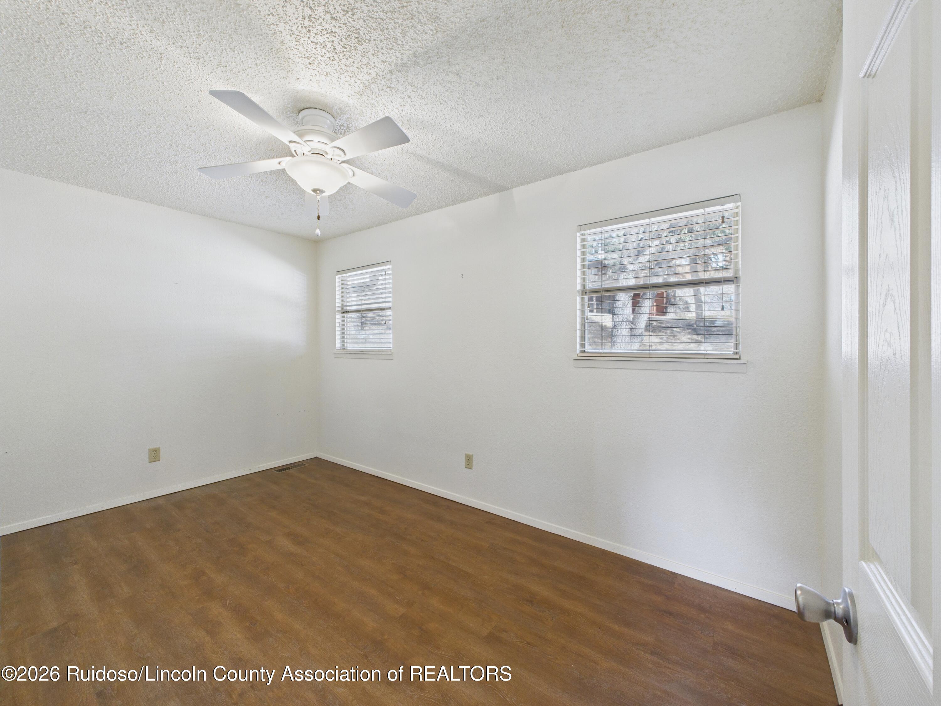 Empty room with brown flooring, two windows with blinds, and a ceiling fan.