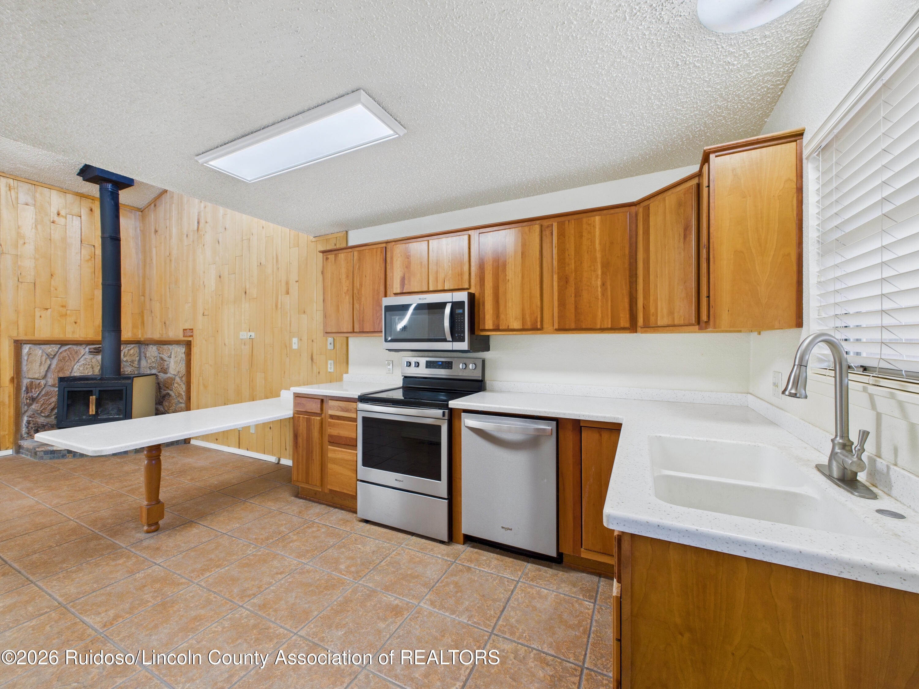 Kitchen with wood cabinetry, stainless steel appliances, a white peninsula, and a wood stove by a stone hearth.