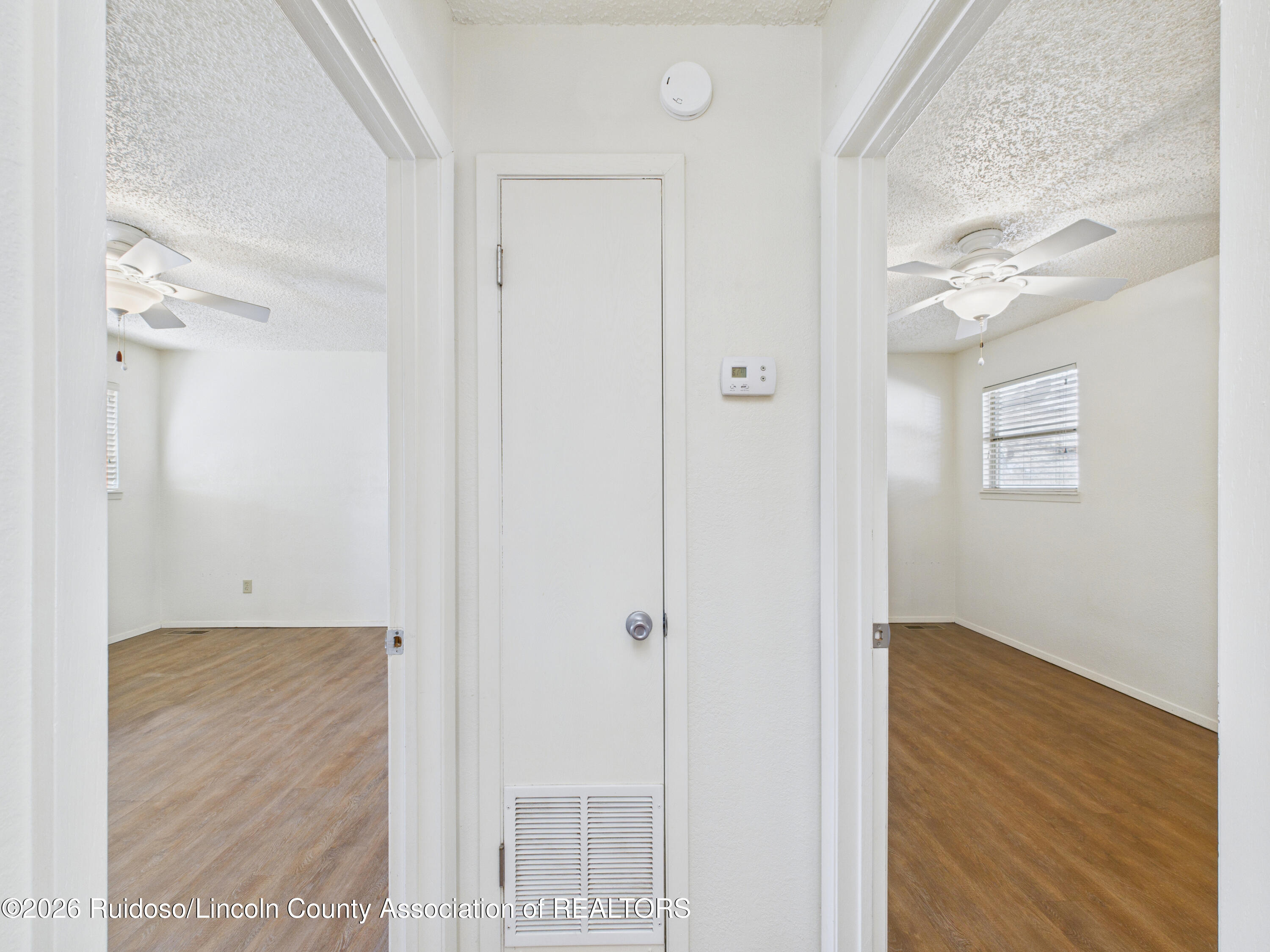 Vacant hallway with a louvered closet door opening onto two rooms with wood-look floors and ceiling fans.