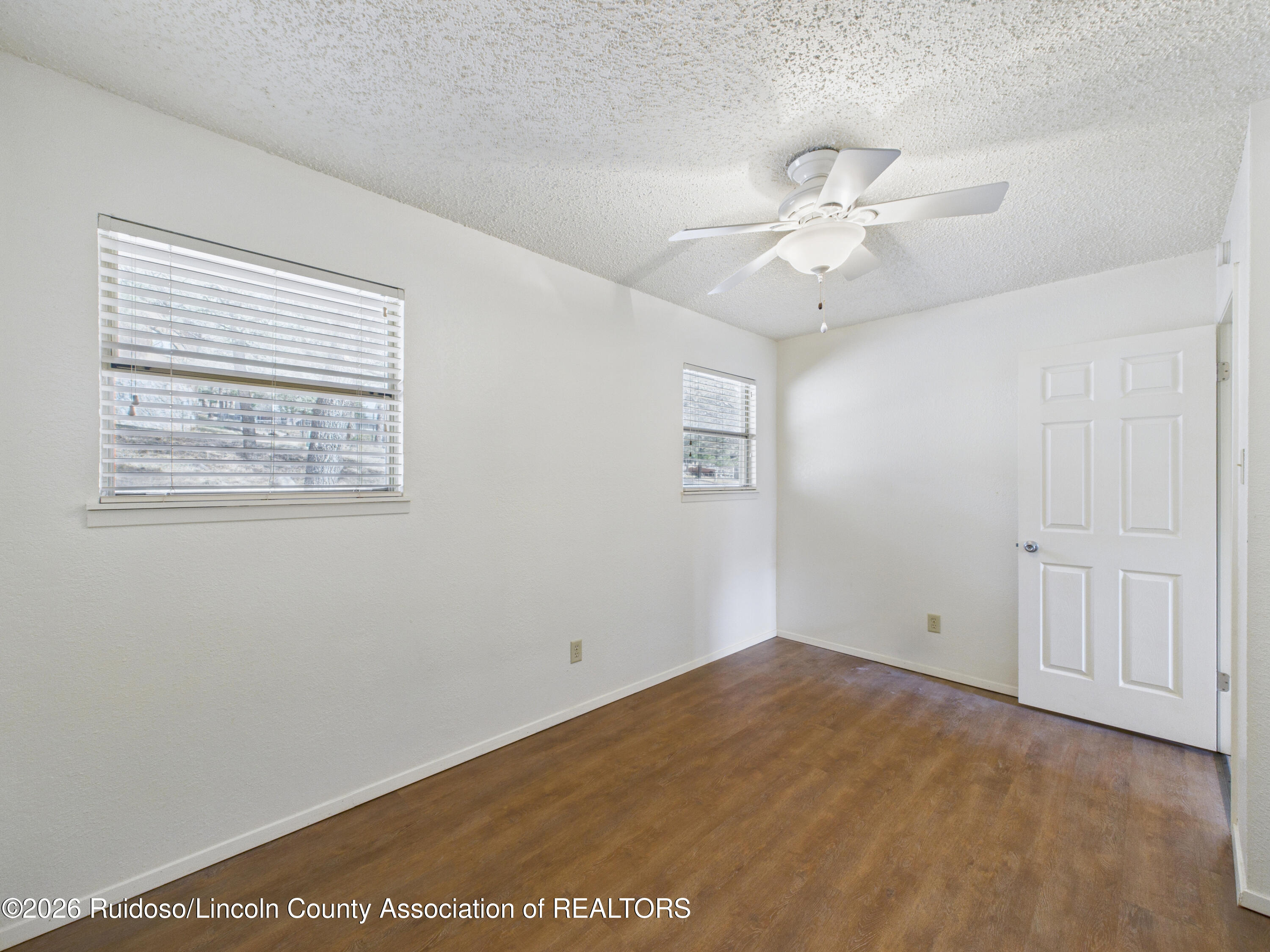 Vacant room with brown wood flooring, two windows with blinds, and a white ceiling fan.