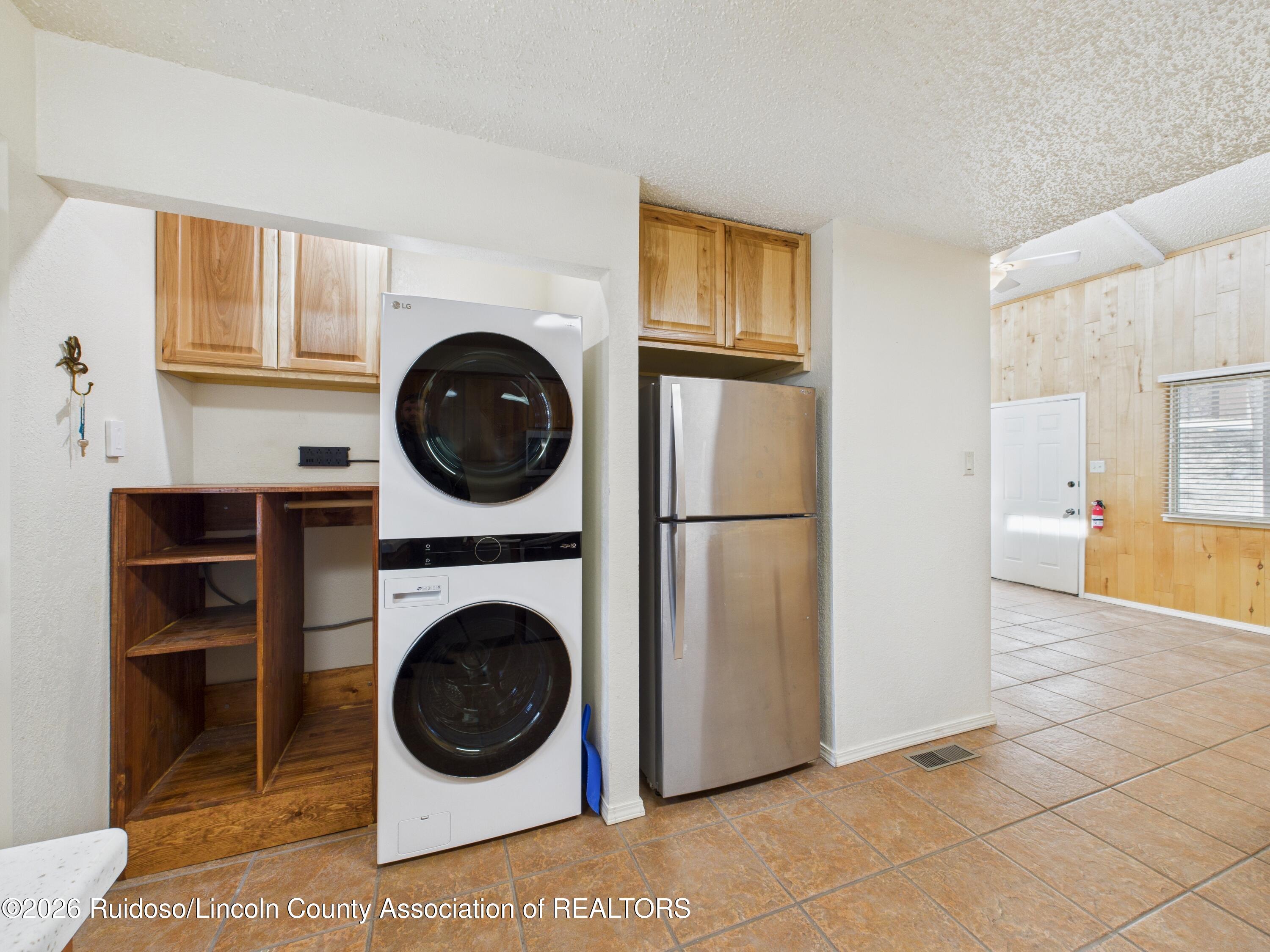 Laundry area with stacked washer and dryer, stainless steel refrigerator, and wood cabinetry.