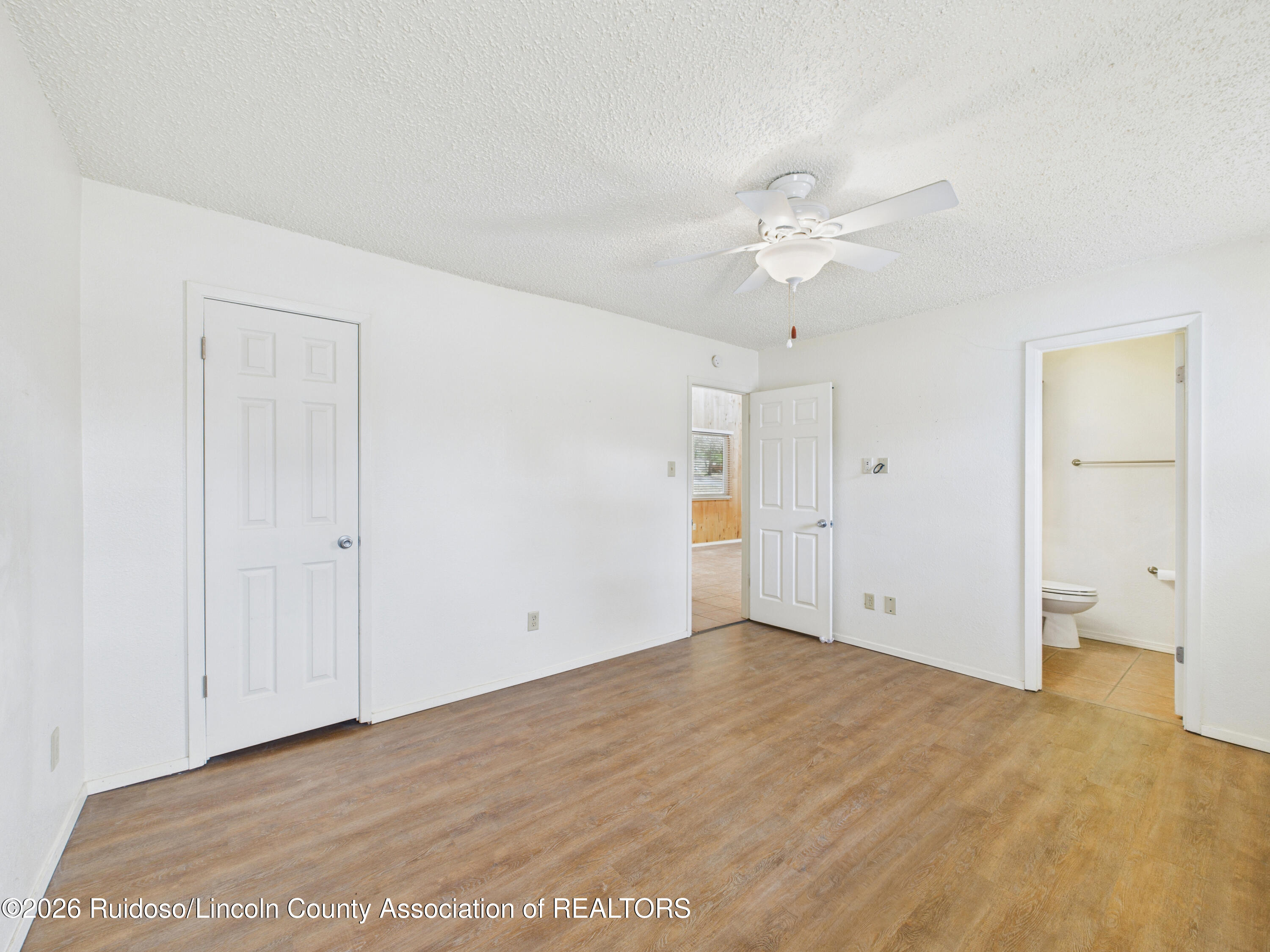 Vacant room with wood-look flooring, a ceiling fan, and an open doorway to a bathroom.