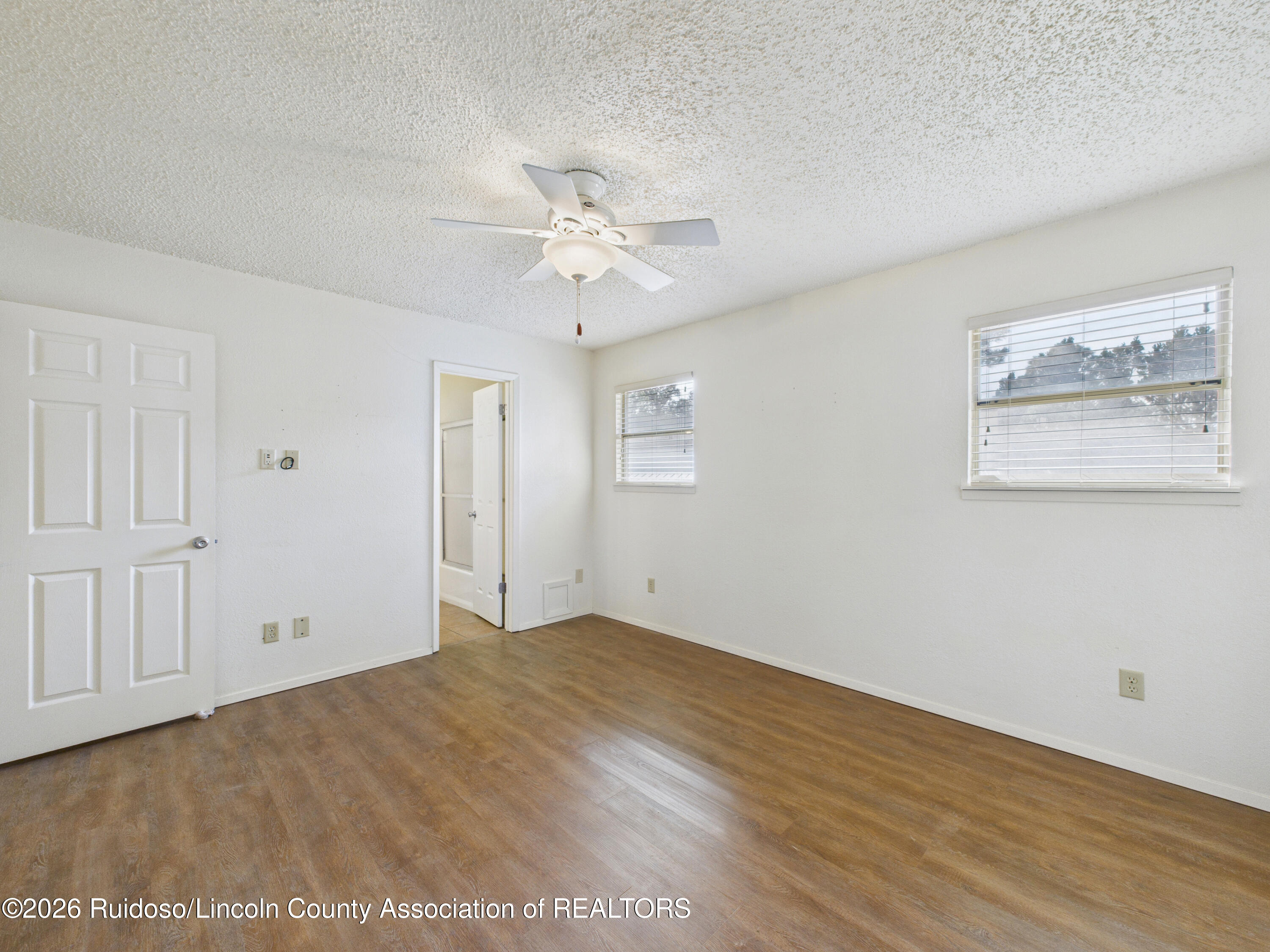 Empty room with brown wood-look flooring, a ceiling fan, and two windows with blinds.