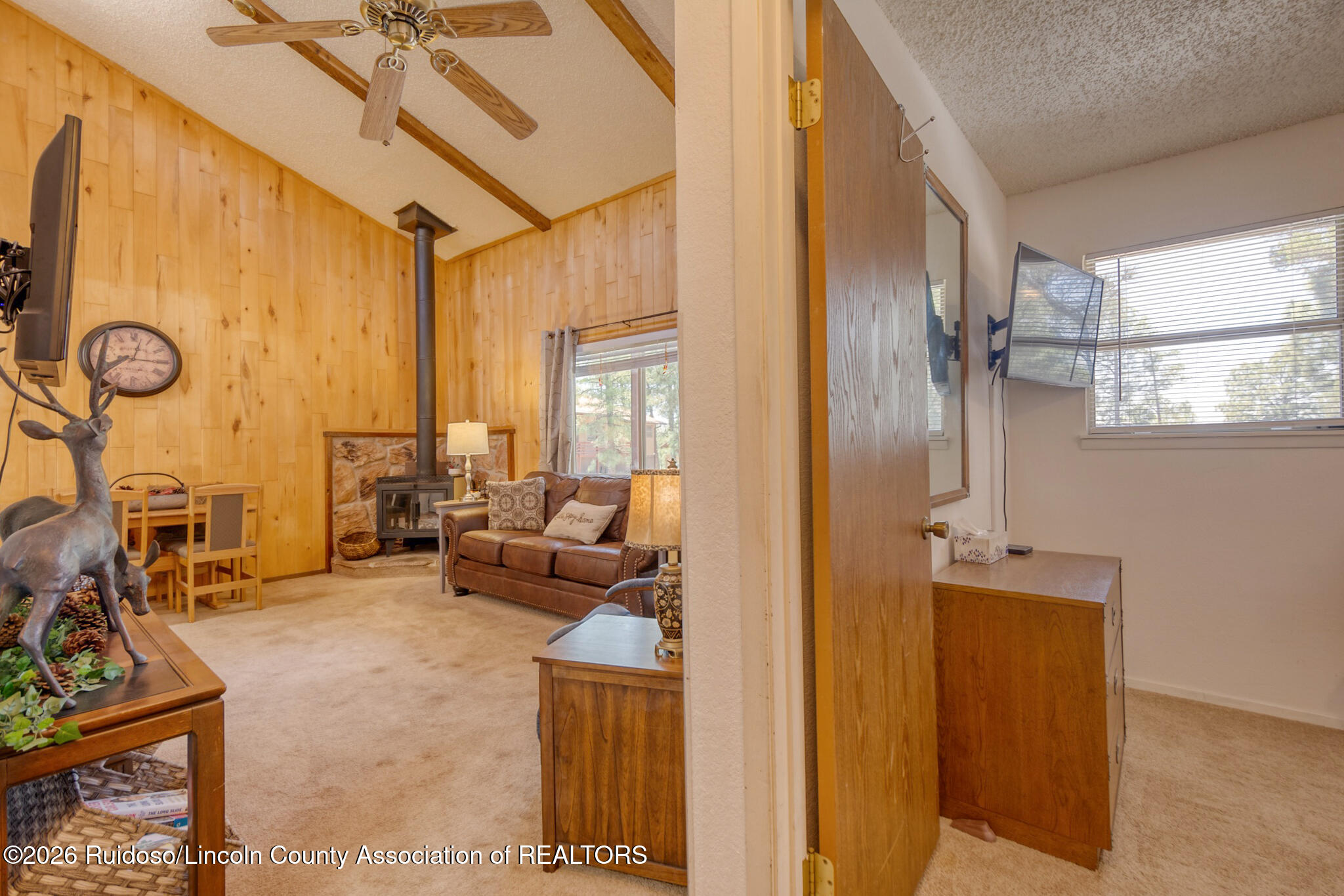Wood-paneled living room with exposed beams, a wood stove, and an adjoining bedroom.