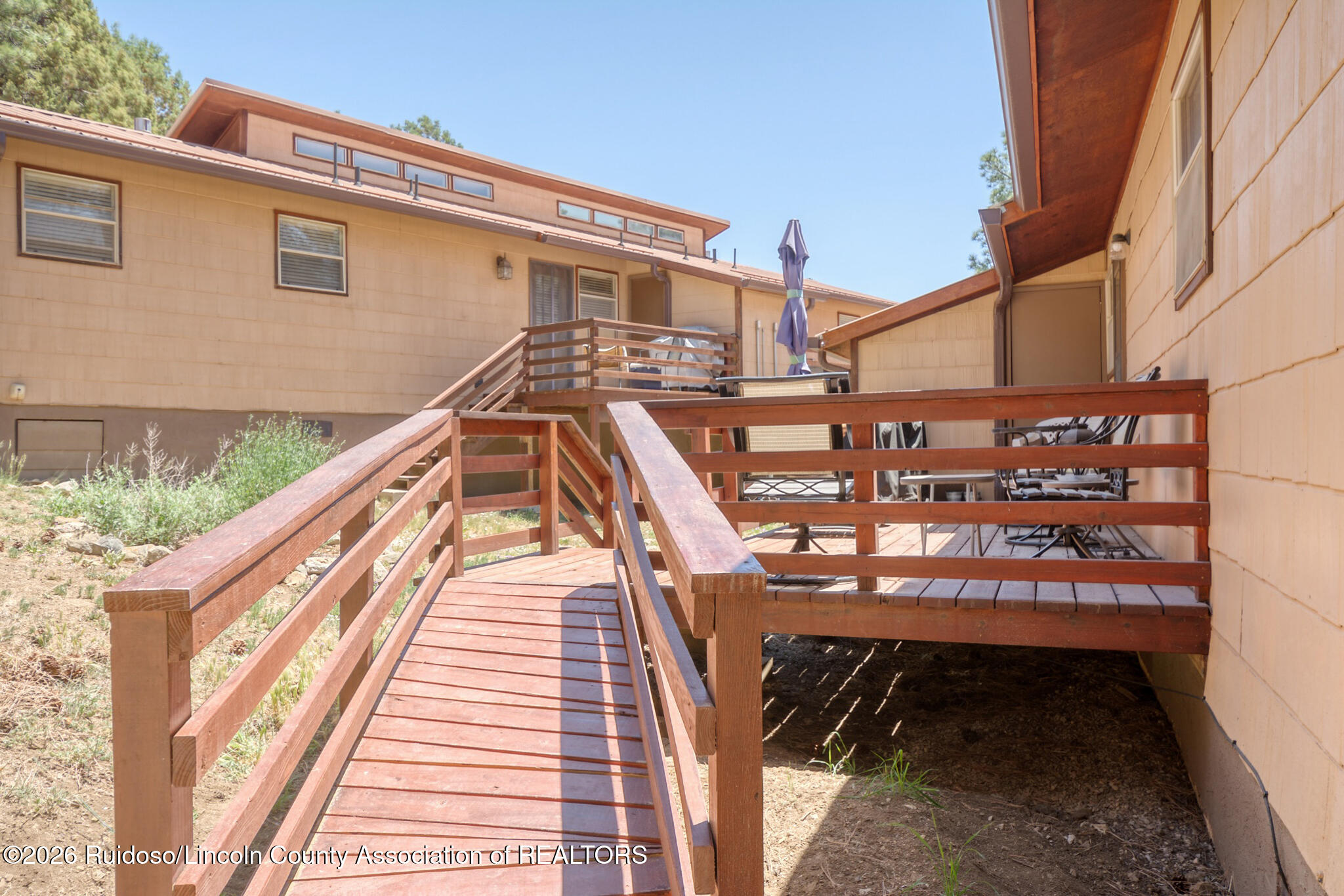 Wooden access ramp leads to a raised deck with patio seating beside a tan-sided home.