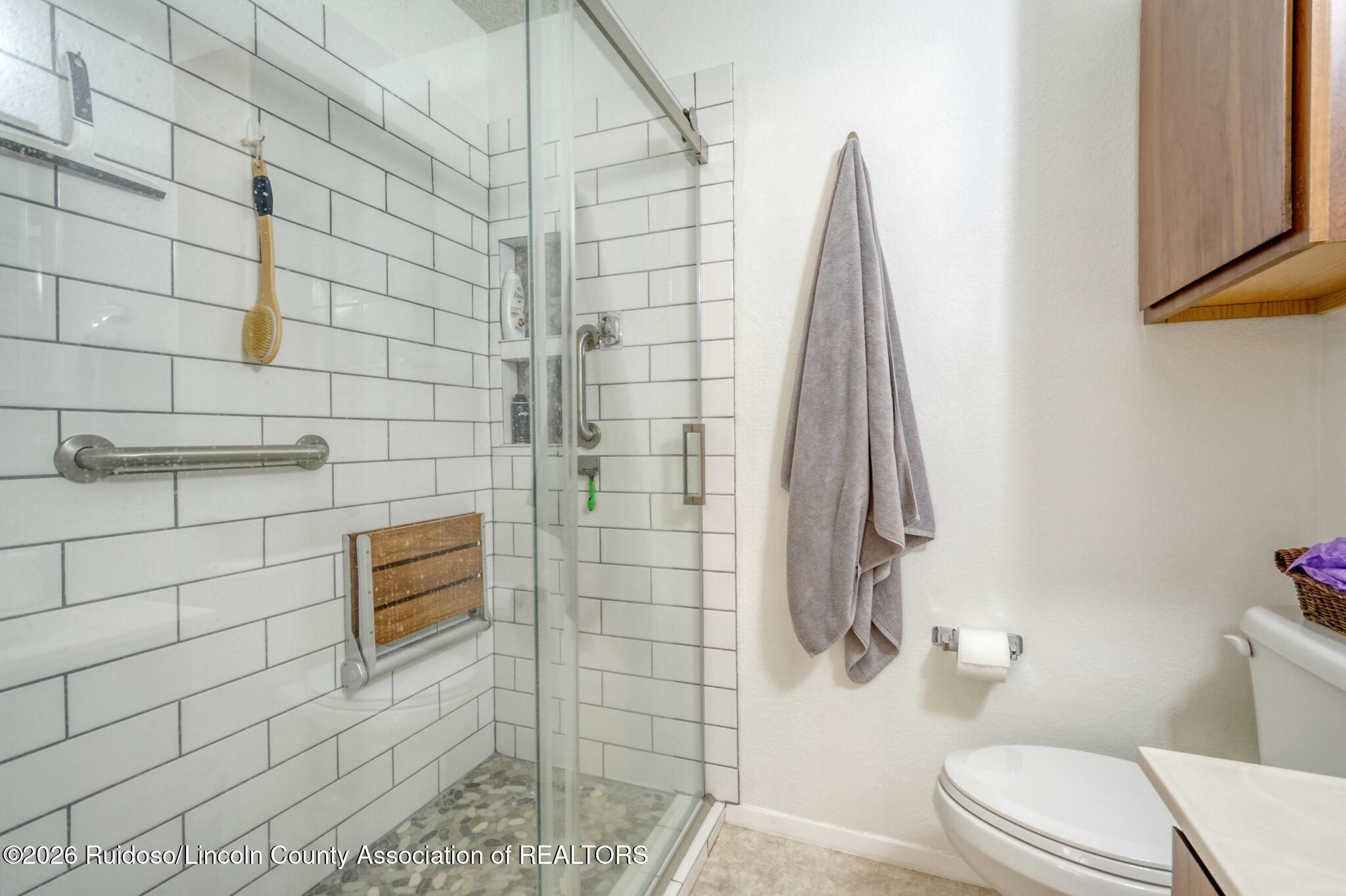 Bathroom with a glass shower, white subway tile, pebble floor, fold-down wooden seat, and grab bars.
