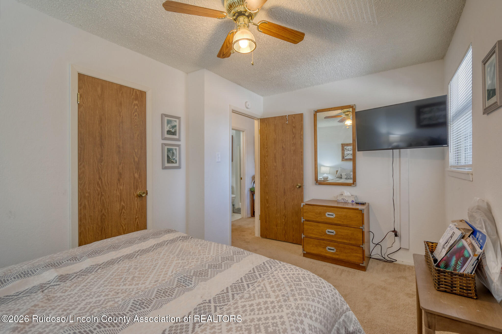 Bedroom with patterned bedspread, wooden dresser, ceiling fan, and wall-mounted television.