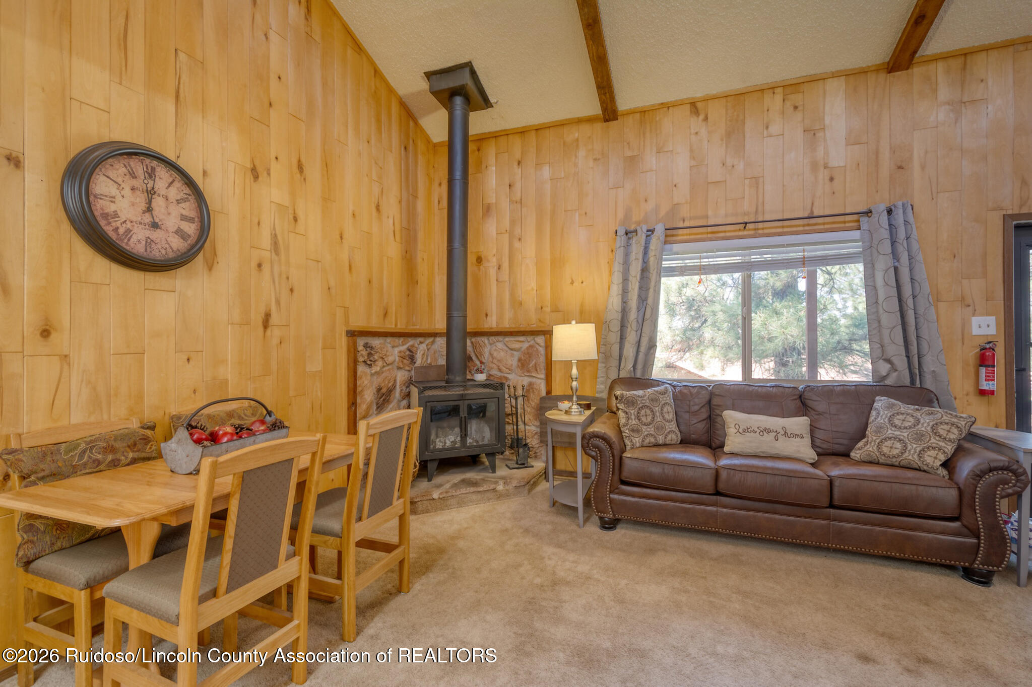 Living and dining area with wood-paneled walls, a wood stove, and a leather sofa beside a large window.