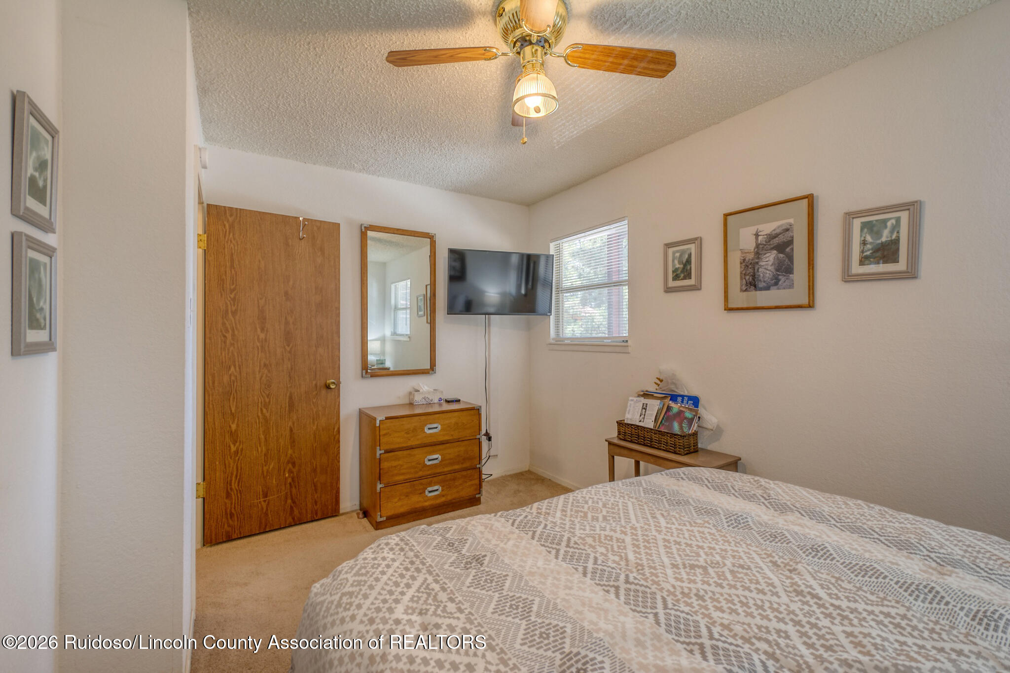 Bedroom with patterned bedspread, wooden dresser, wall-mounted television, and ceiling fan.