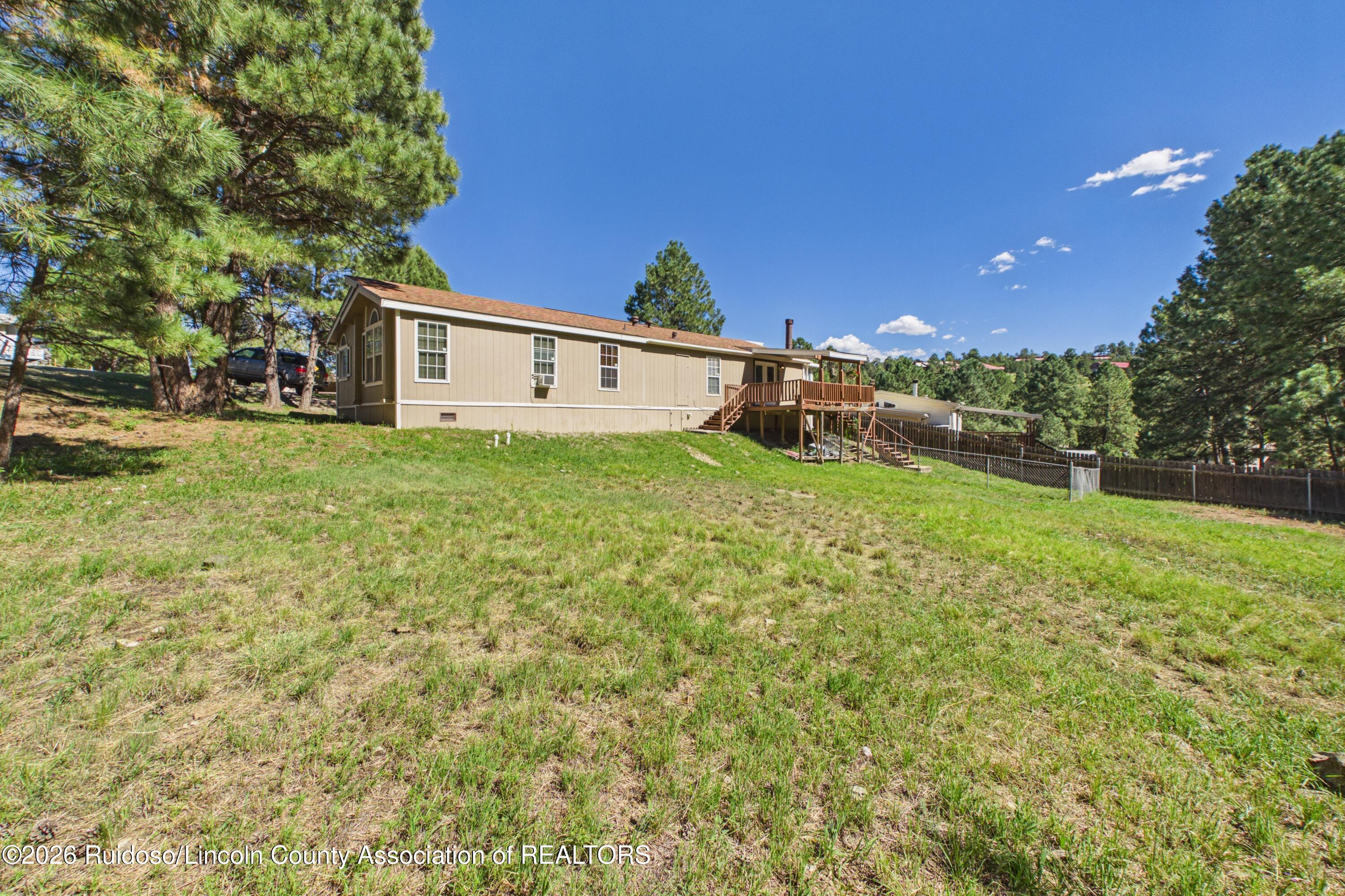 Tan single-story home with a raised wooden deck and stairs on a grassy, tree-lined lot.