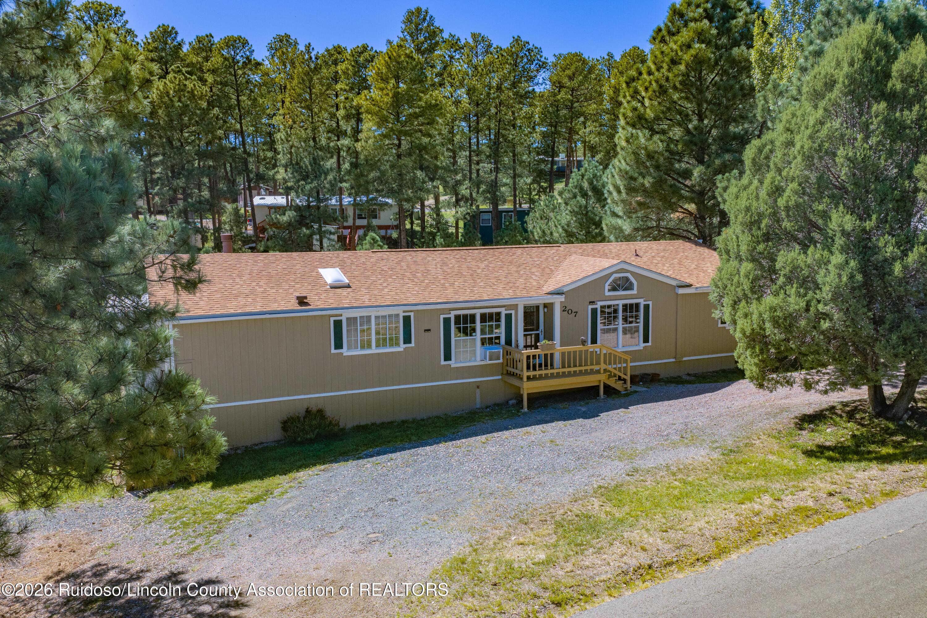 Tan home with a wooden front deck, gravel driveway, and tall pine trees.