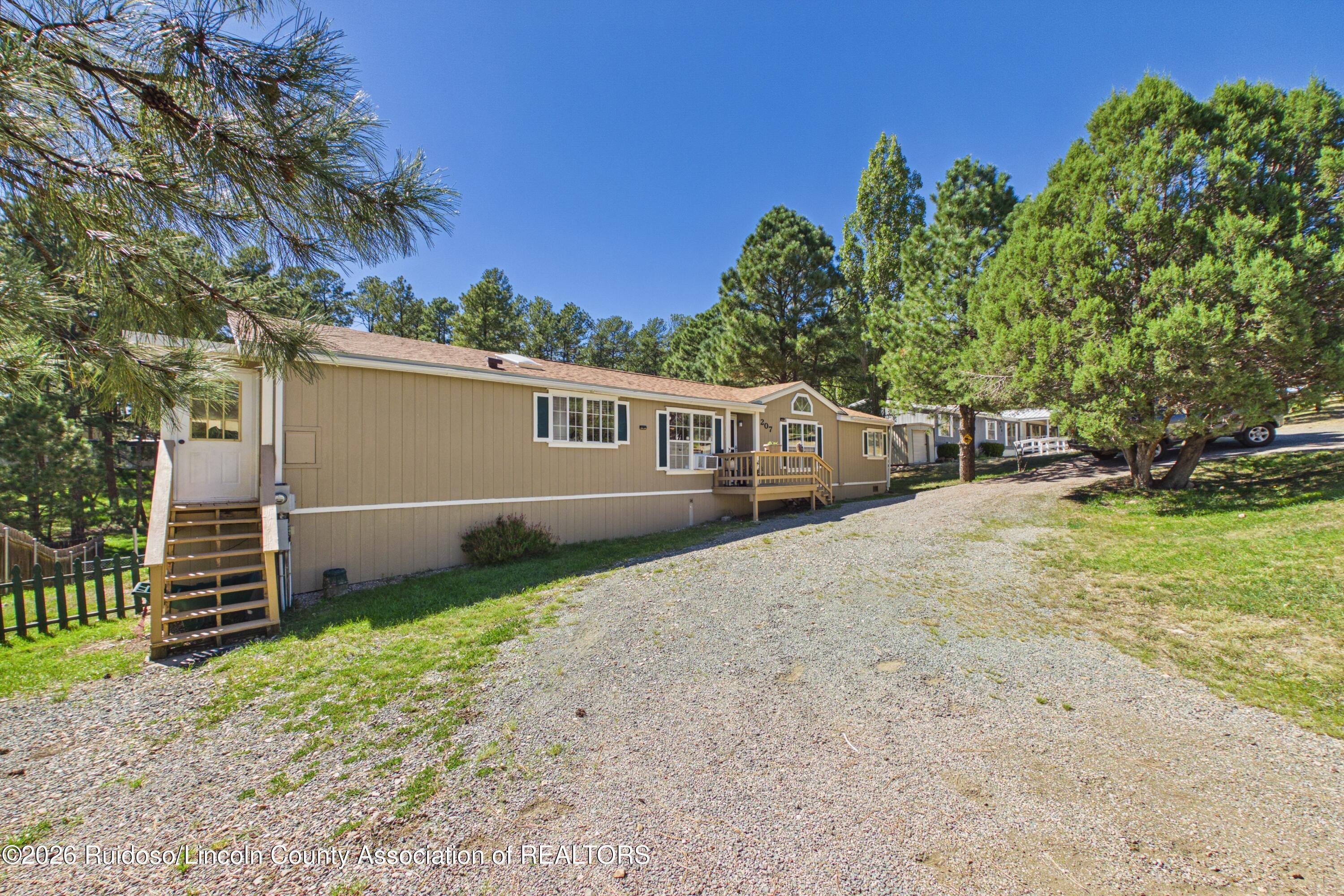Tan single-story home with two wooden stairways beside a gravel driveway, surrounded by pine trees.