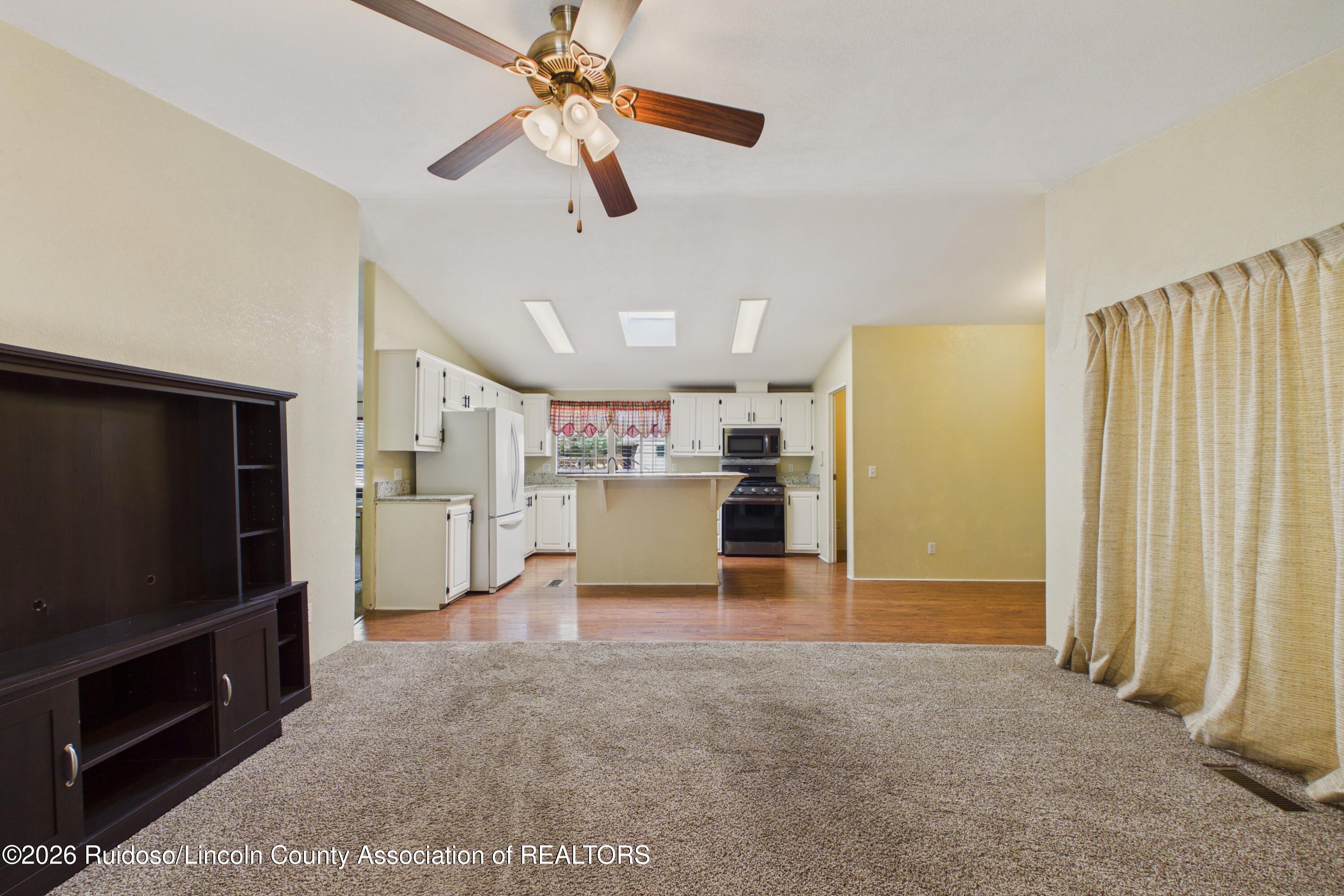 Living room with beige carpet and dark media cabinet, open to a kitchen with white cabinets and breakfast bar.