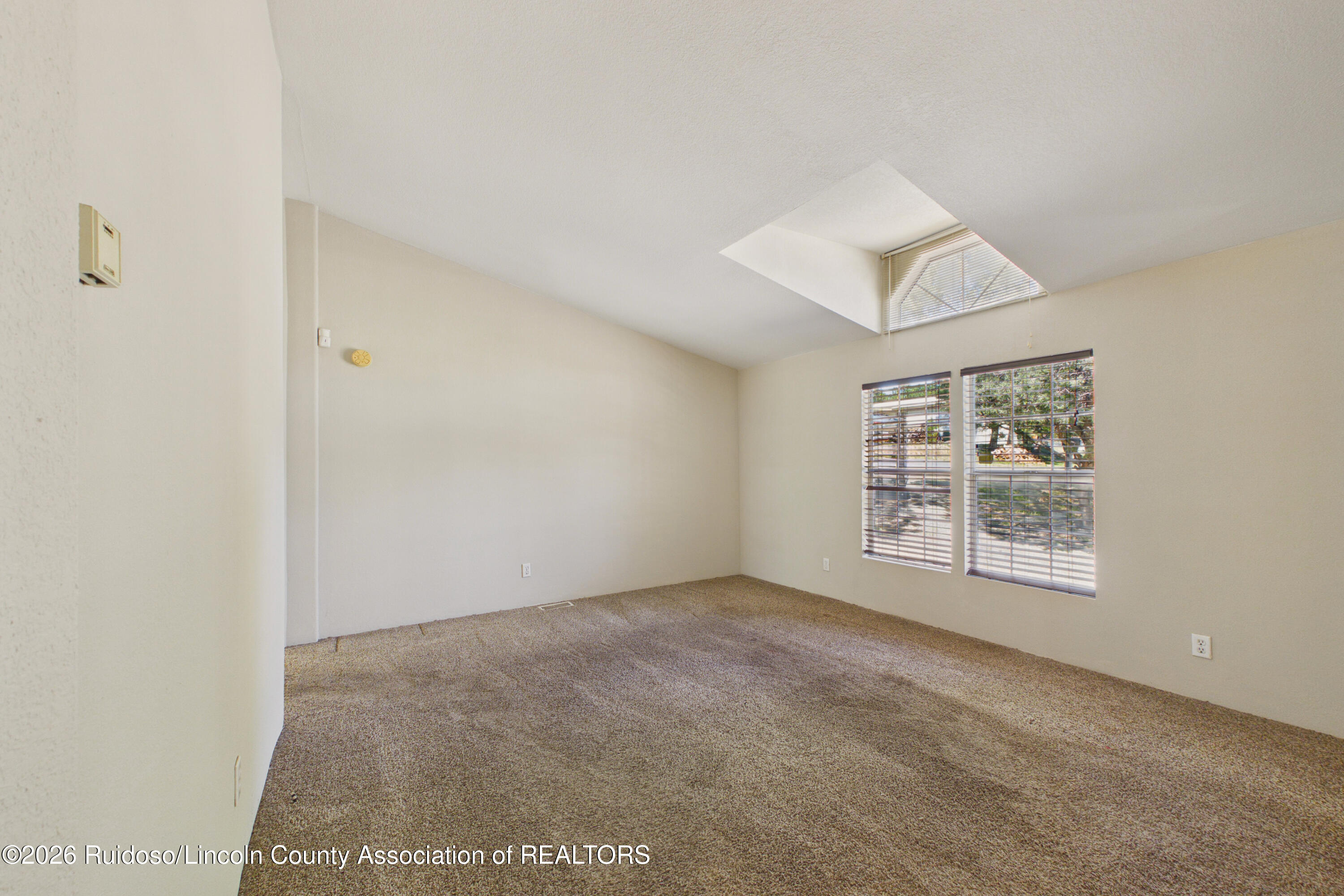 Vacant carpeted room with a vaulted ceiling and large windows with blinds.