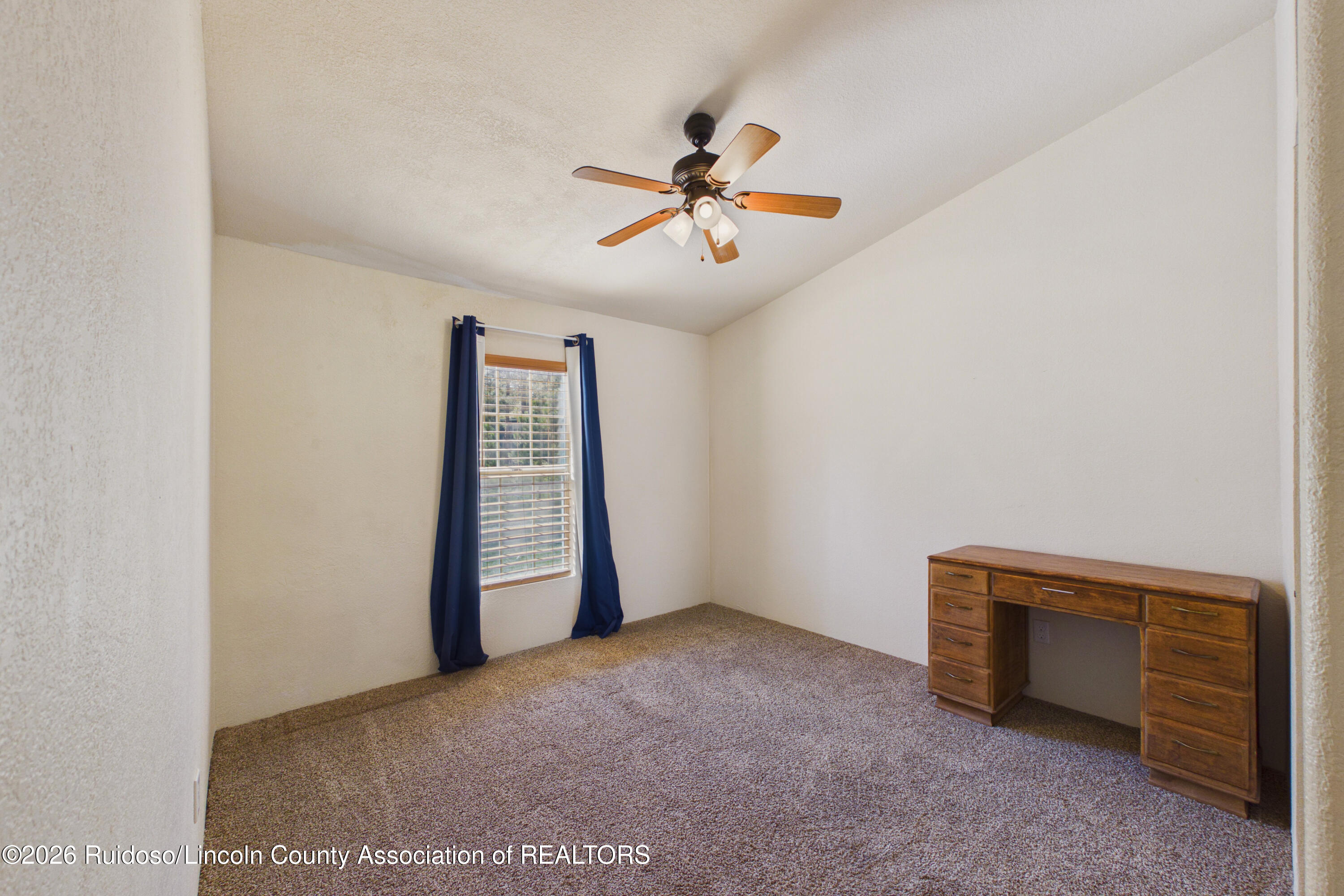 Carpeted room with a wooden desk, blue curtains, and a ceiling fan with light.