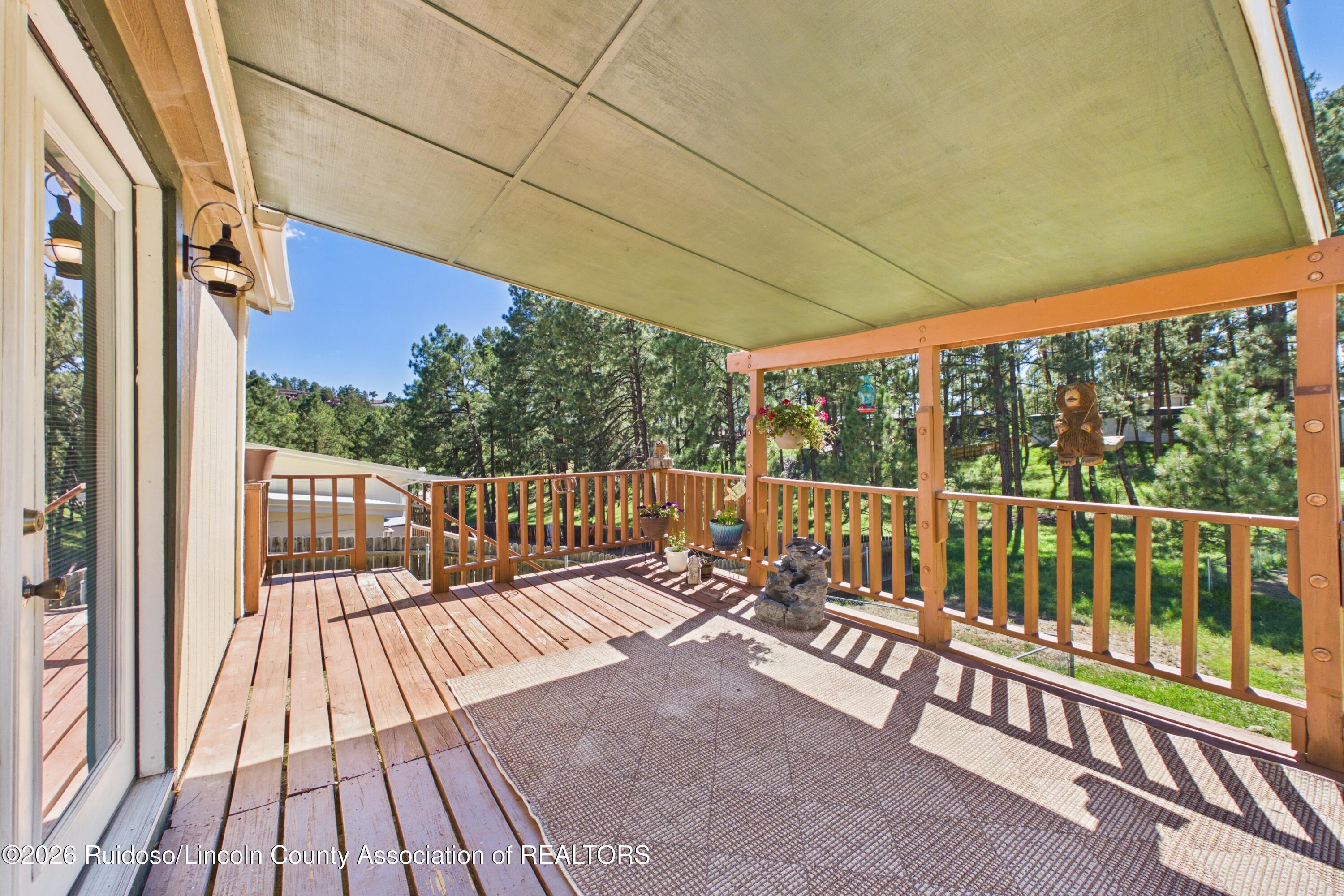 Covered wooden deck with an outdoor rug and potted flowers overlooking pine trees.