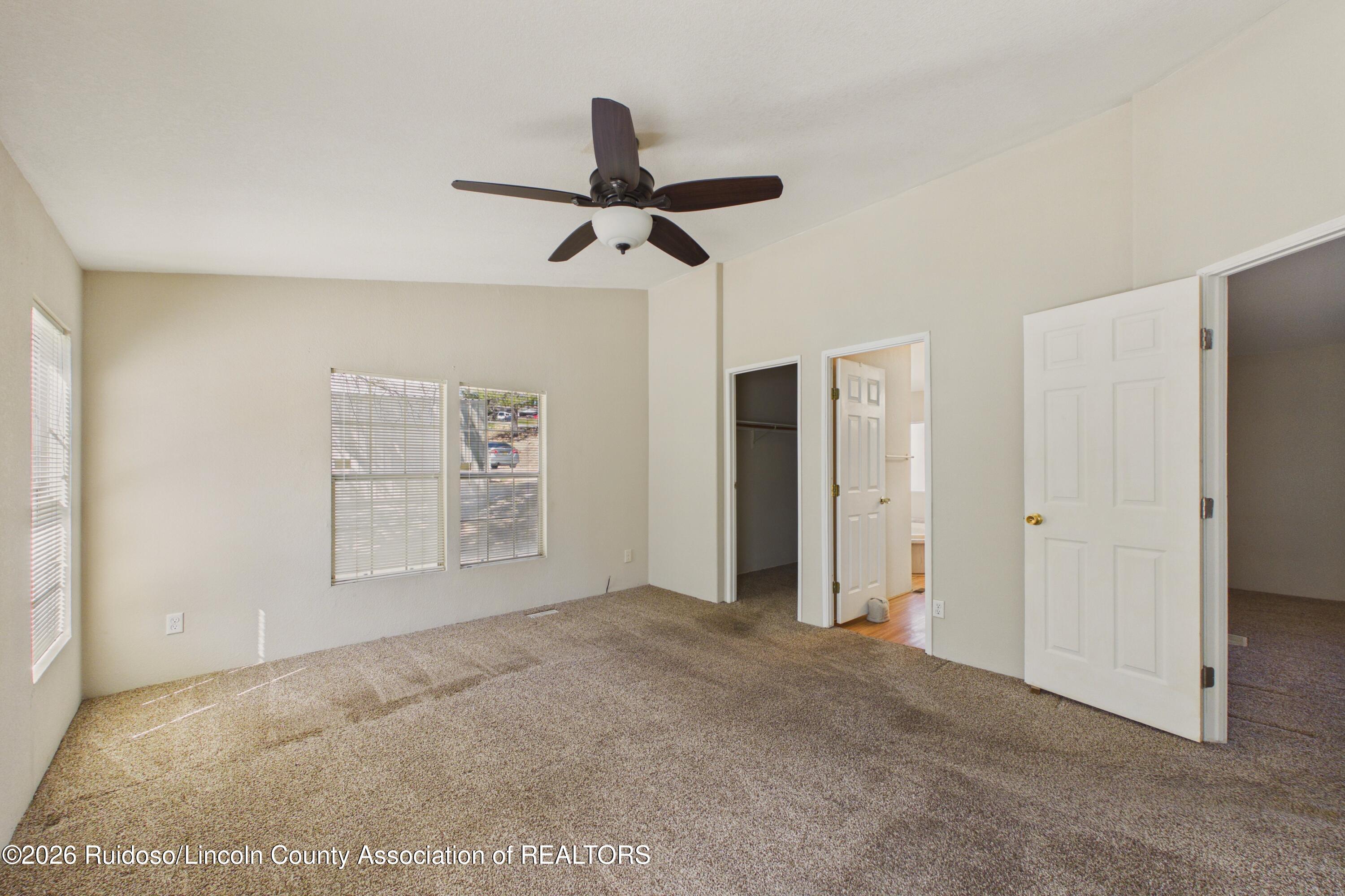 Vacant carpeted bedroom with two windows, a ceiling fan, and open closet and bathroom doors.