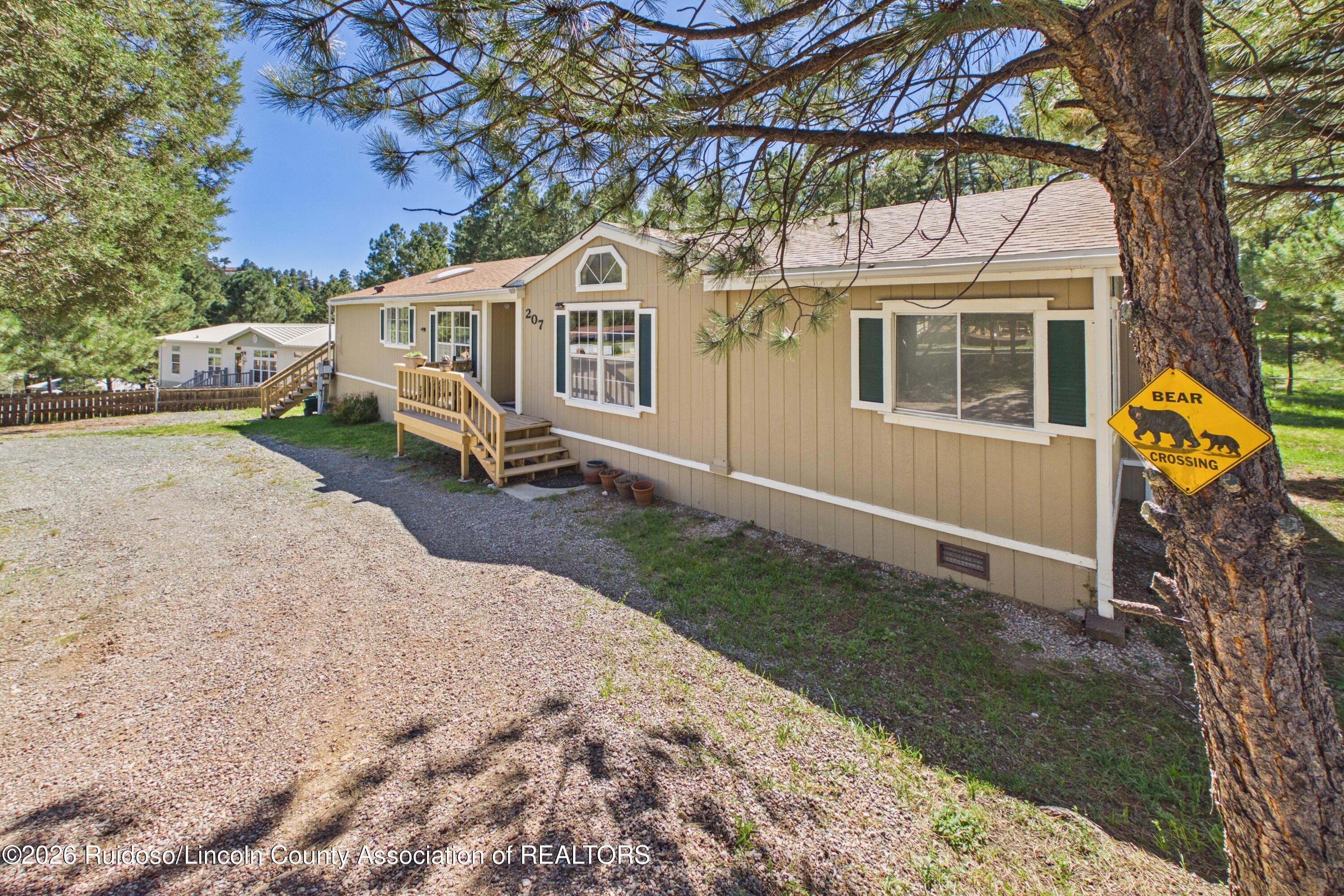 Tan home with wooden entry steps beneath pine trees; gravel driveway and bear crossing sign.