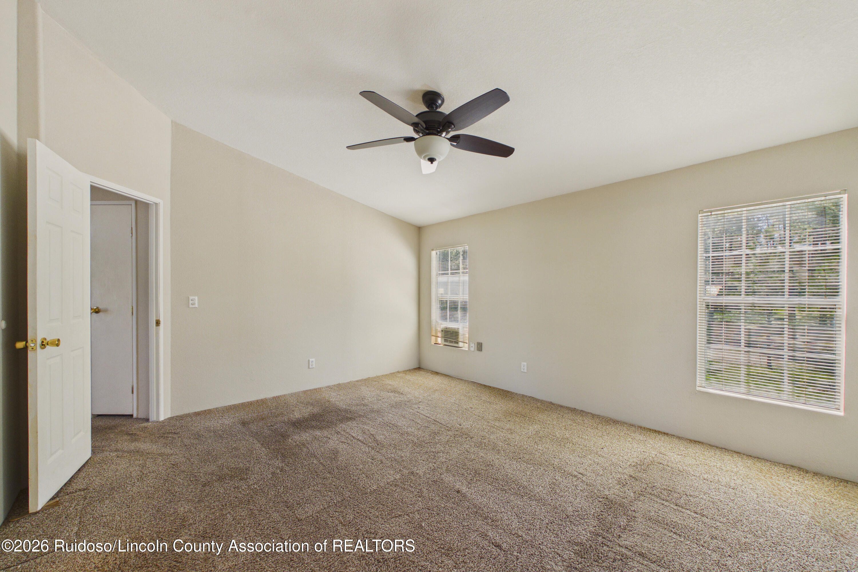 Empty bedroom with beige carpet, a dark ceiling fan, and two windows with blinds.
