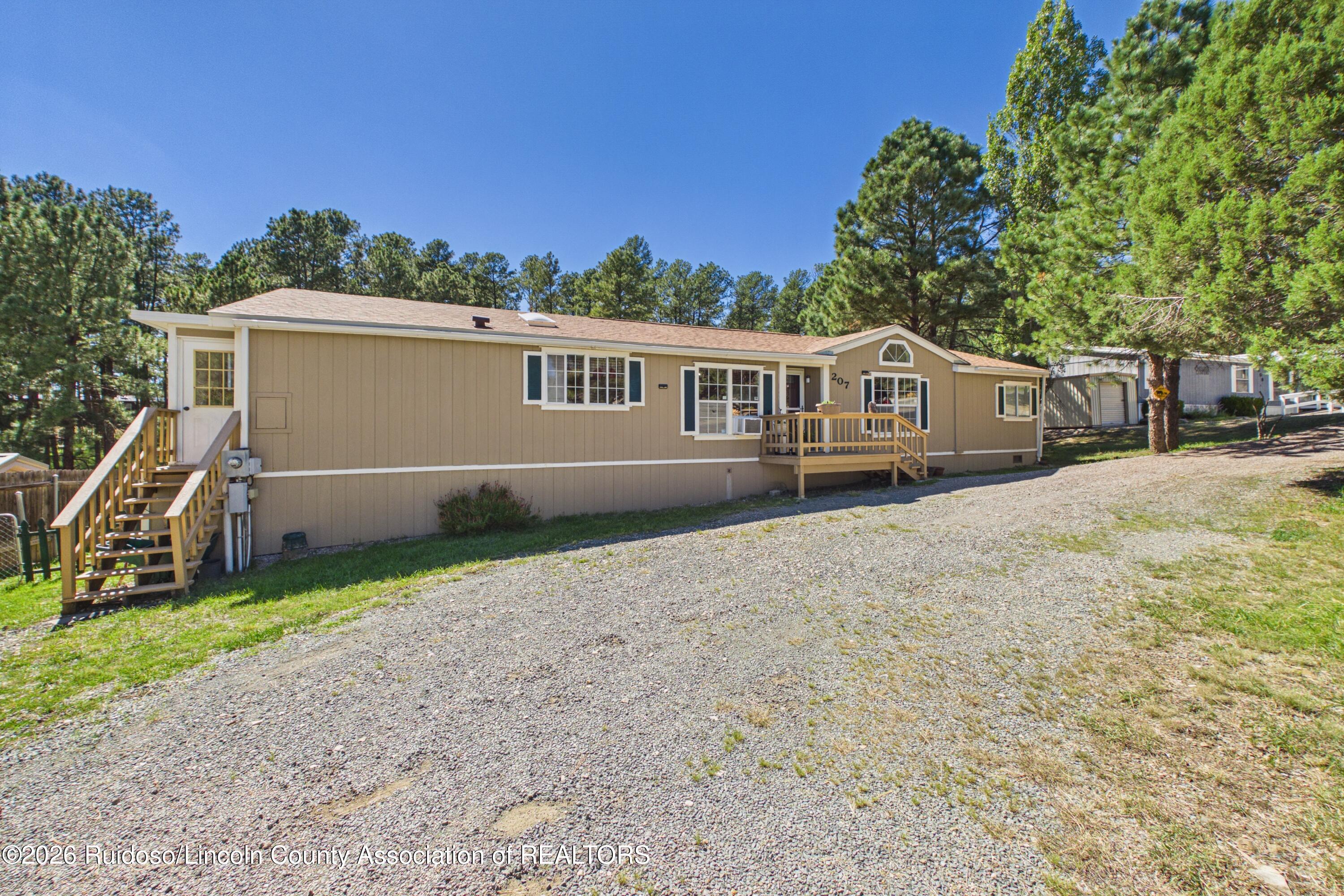 Tan single-story home with wooden entry decks and stairs, a gravel driveway, and pine trees.
