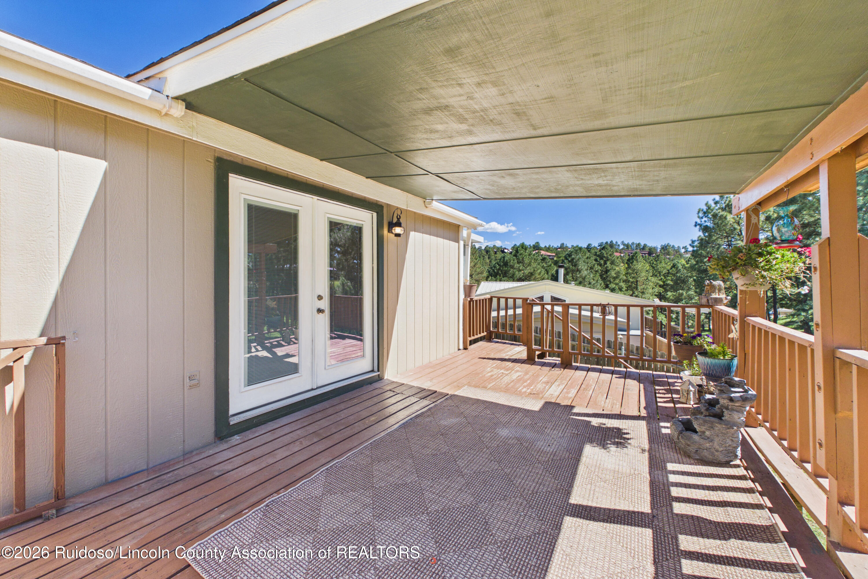 Covered wooden deck with French doors, potted plants, and a view of pine trees.