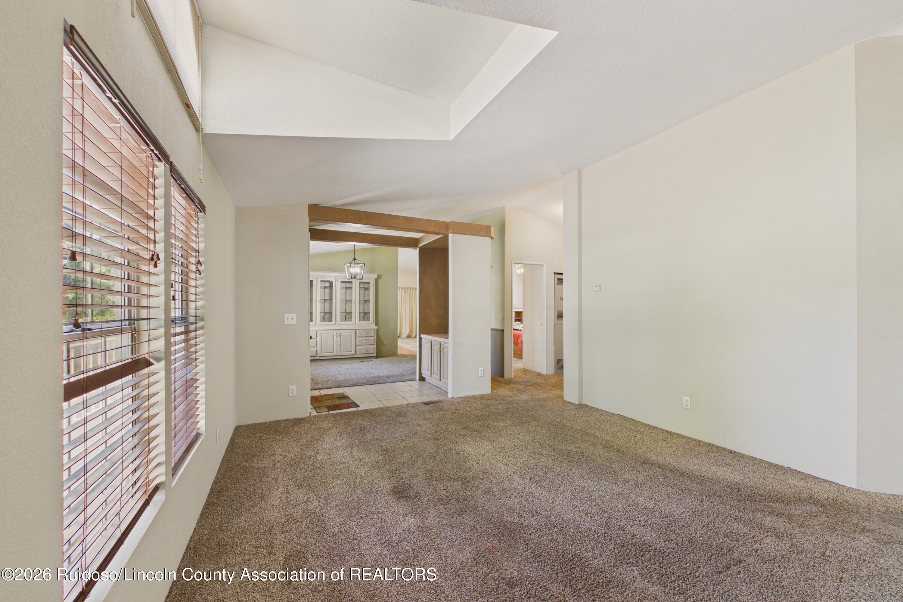Carpeted living area with large windows, blinds, and an opening to an adjoining room.
