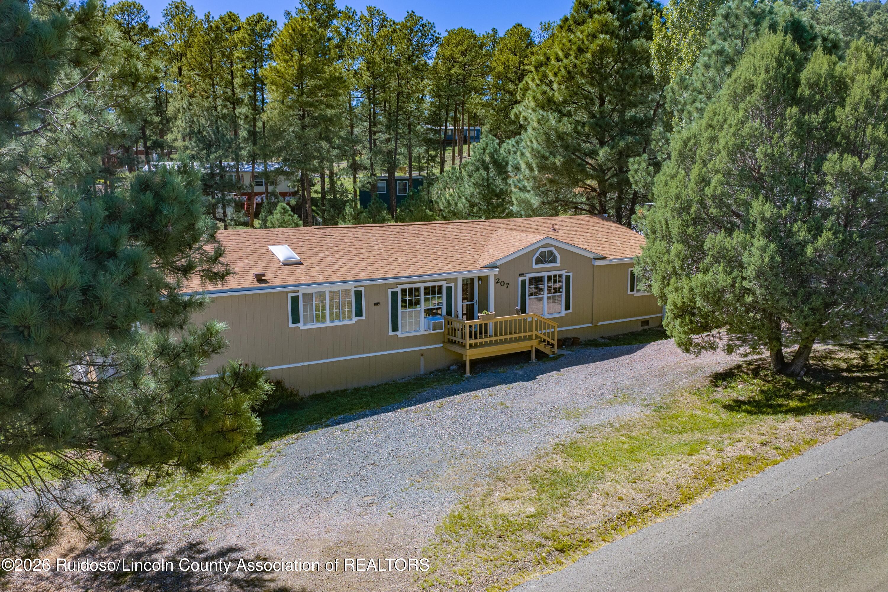Tan single-story home with a small wooden porch, gravel driveway, and surrounding pine trees.