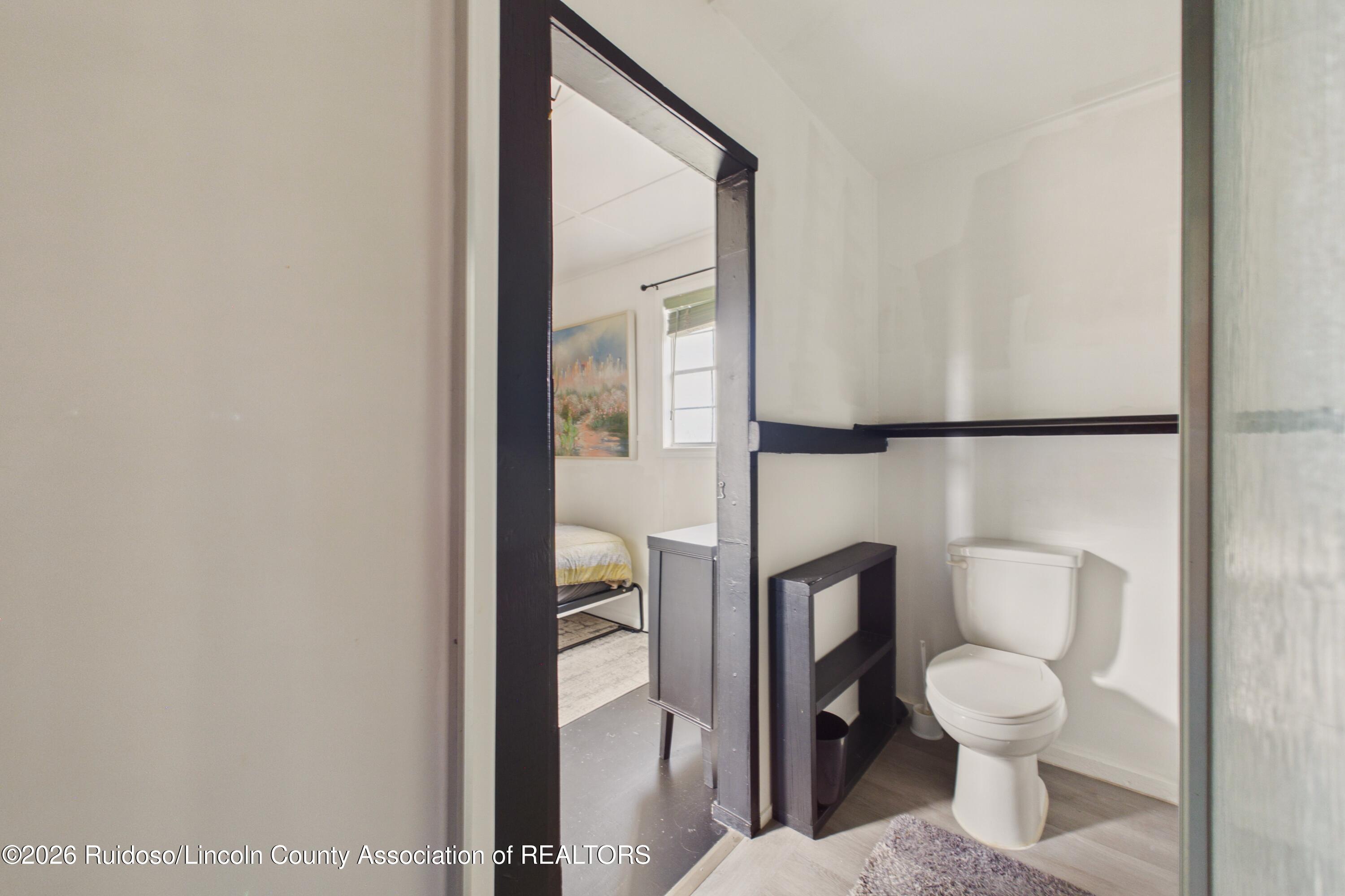 Bathroom with a white toilet and dark shelving, looking through a doorway toward an adjoining bedroom.
