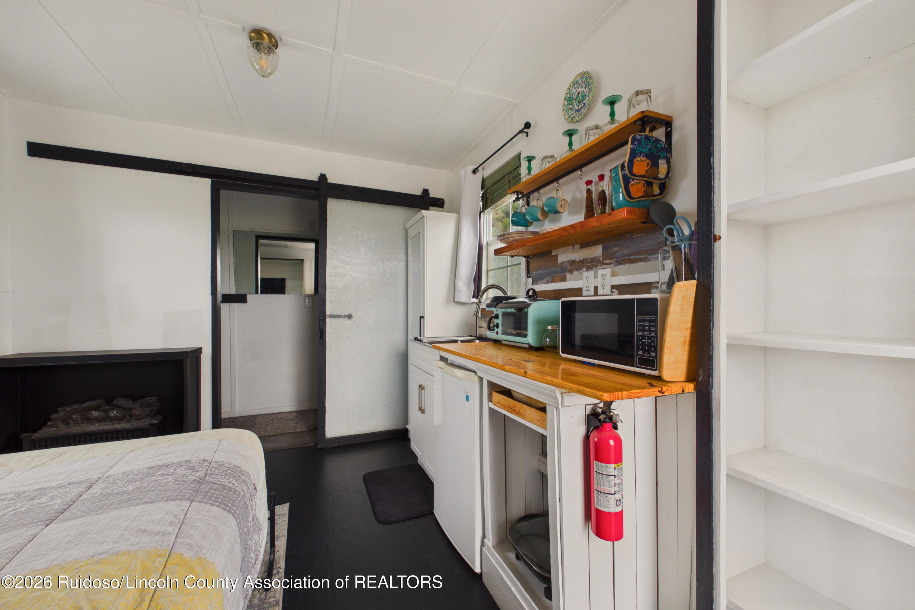 Compact kitchenette with wooden counters and open shelves, beside a bed and dark fireplace.