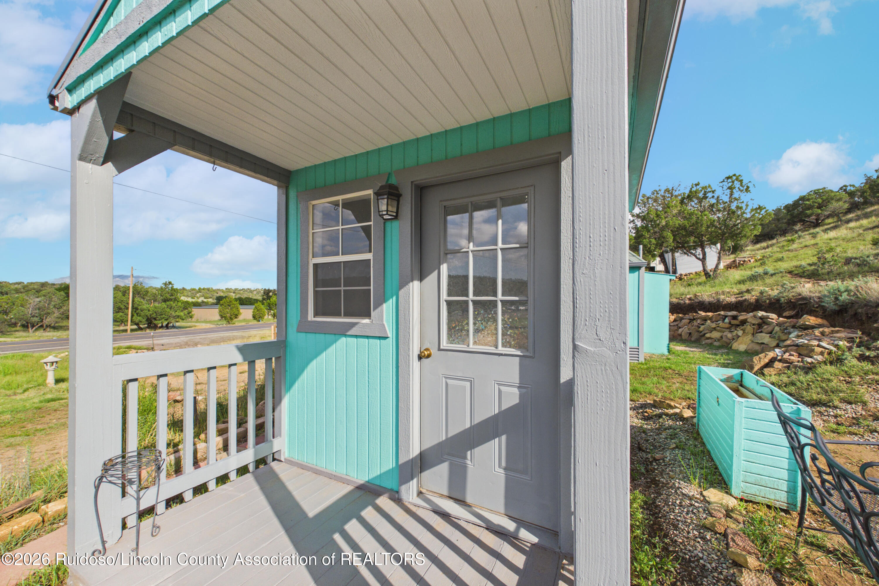 Covered porch with turquoise siding, gray glass-panel door, and views toward the road and surrounding greenery.