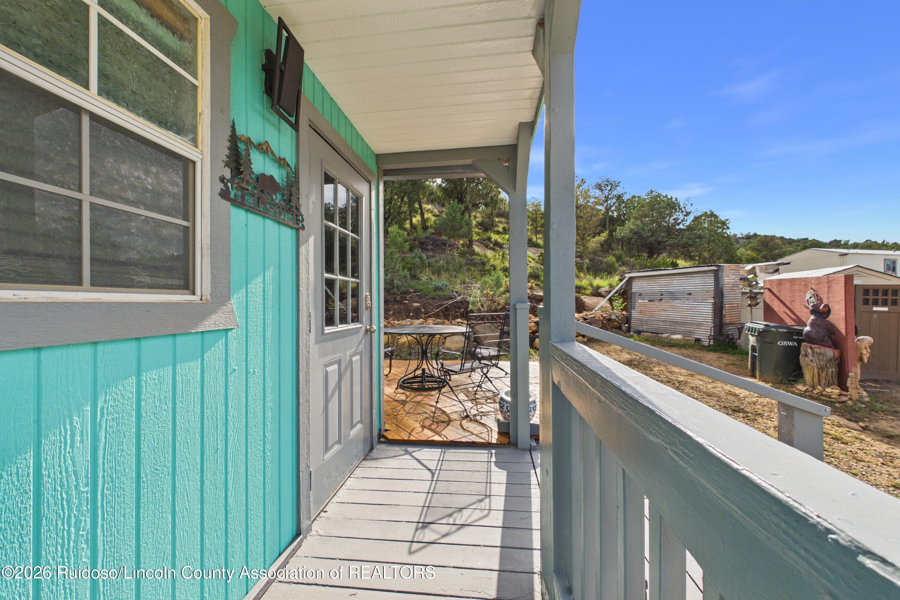 Covered porch beside a turquoise building, with metal patio furniture and sheds in the wooded yard.