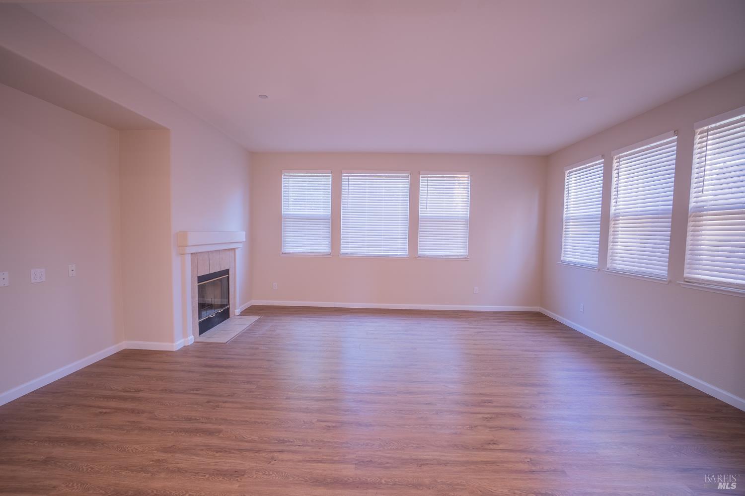Vacant living room with a fireplace, wood-look flooring, and multiple windows with blinds.