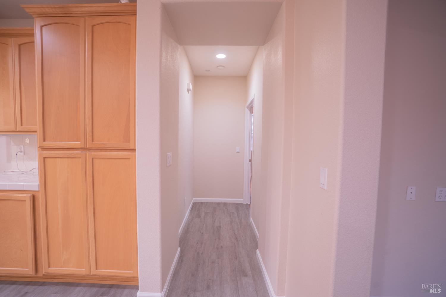 Interior hallway beside honey-colored kitchen cabinets, with gray wood-look flooring and recessed ceiling lights.