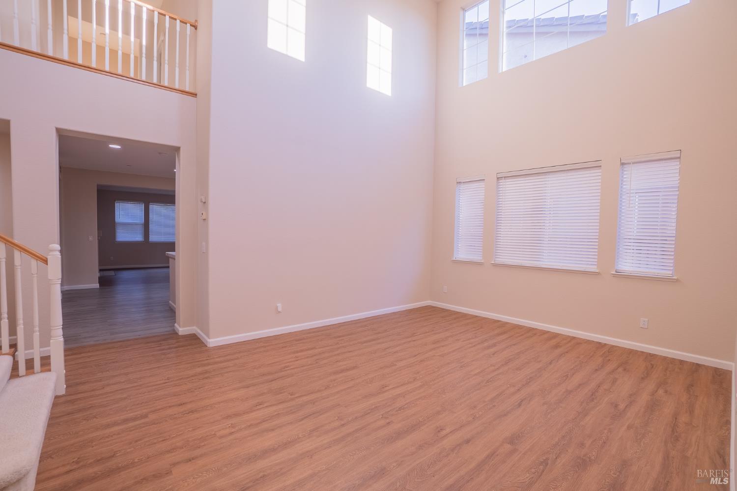 Empty two-story living room with wood-look flooring, tall windows, and an open staircase.
