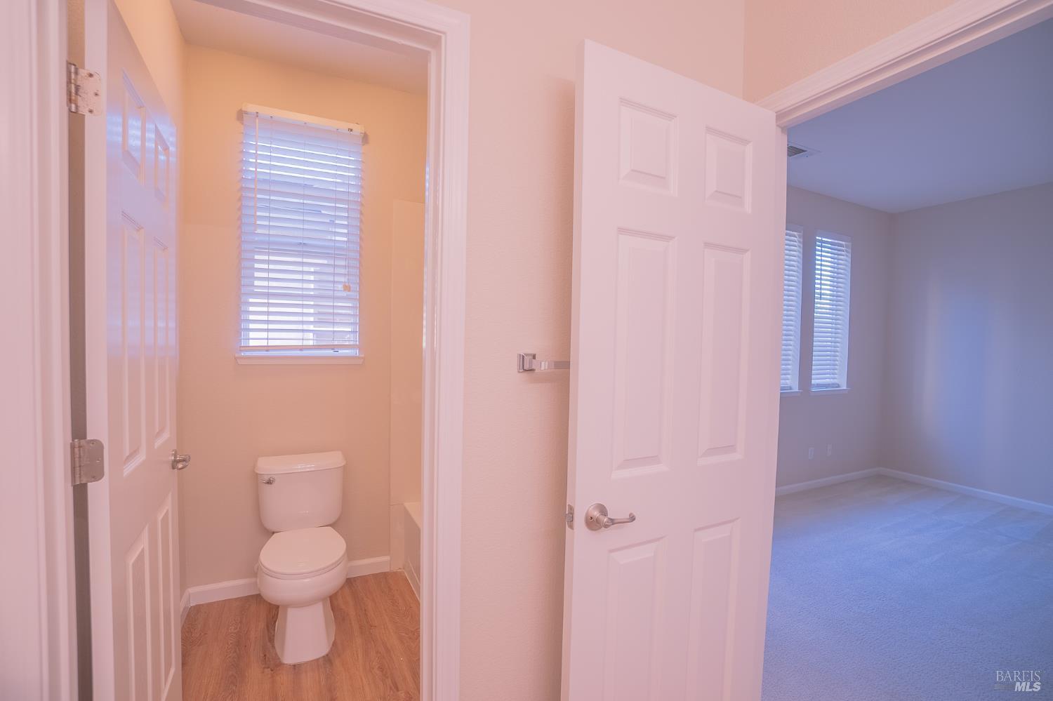 Bathroom with a toilet and wood-look flooring, opening onto an empty carpeted room.