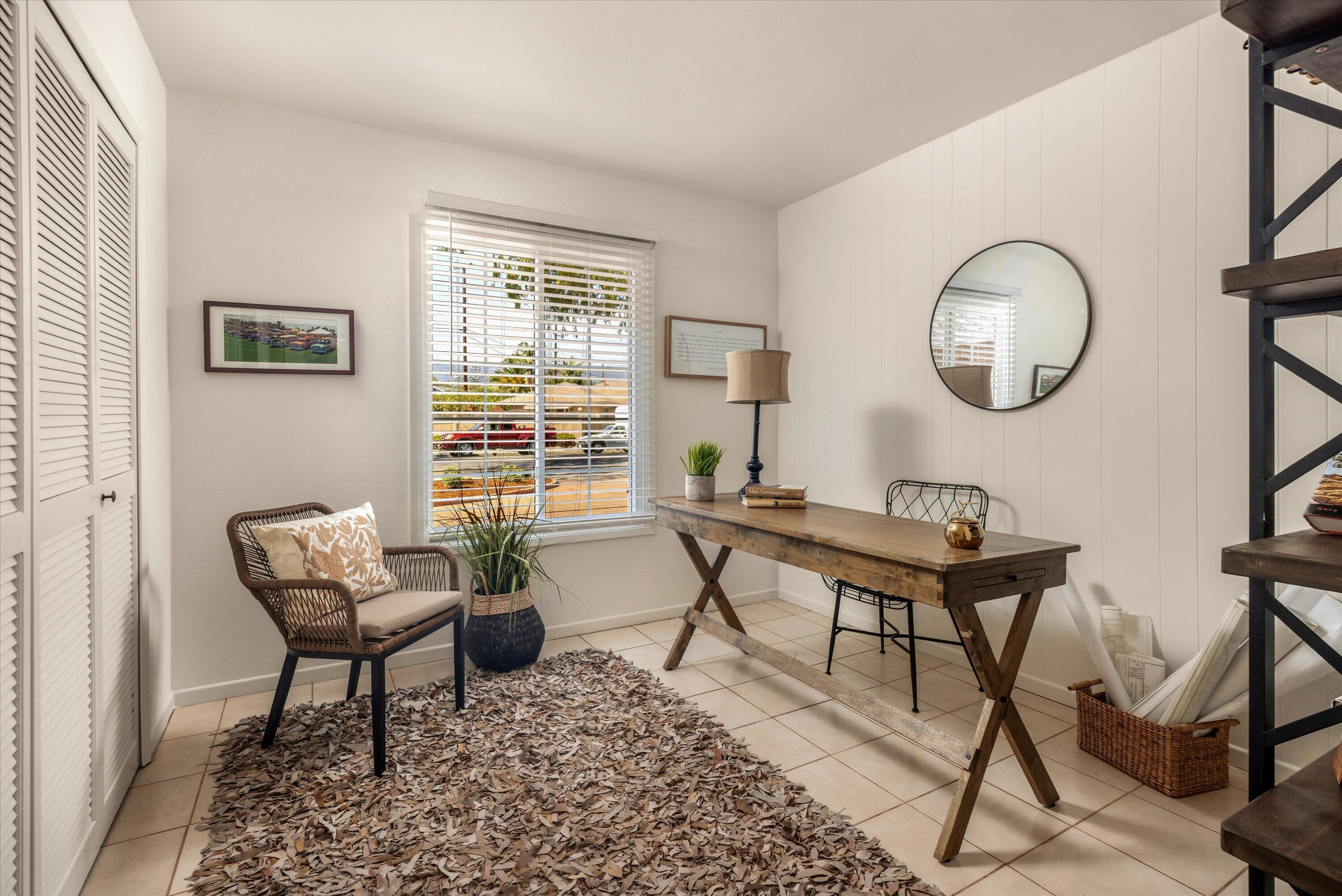 Home office with a rustic wooden desk, woven armchair, and large window with white blinds.