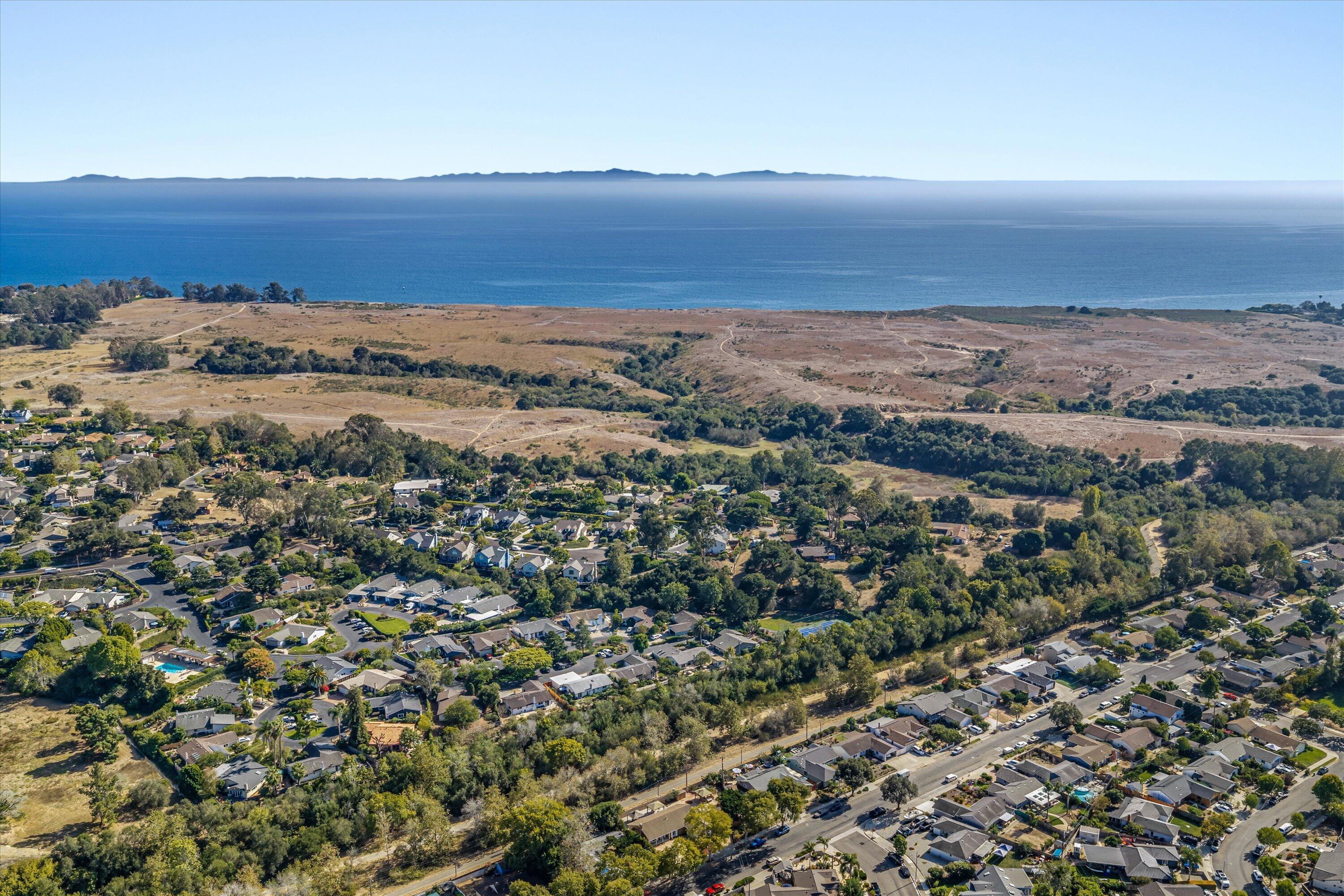 Aerial view of a coastal neighborhood, open space, and ocean with distant mountains.