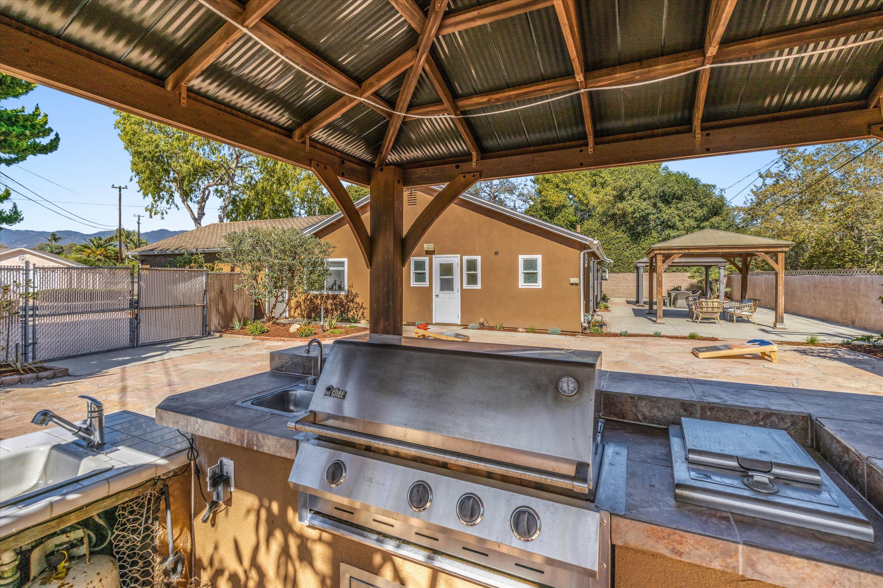 Backyard outdoor kitchen with a stainless-steel grill beneath a timber pavilion and a gazebo beyond.