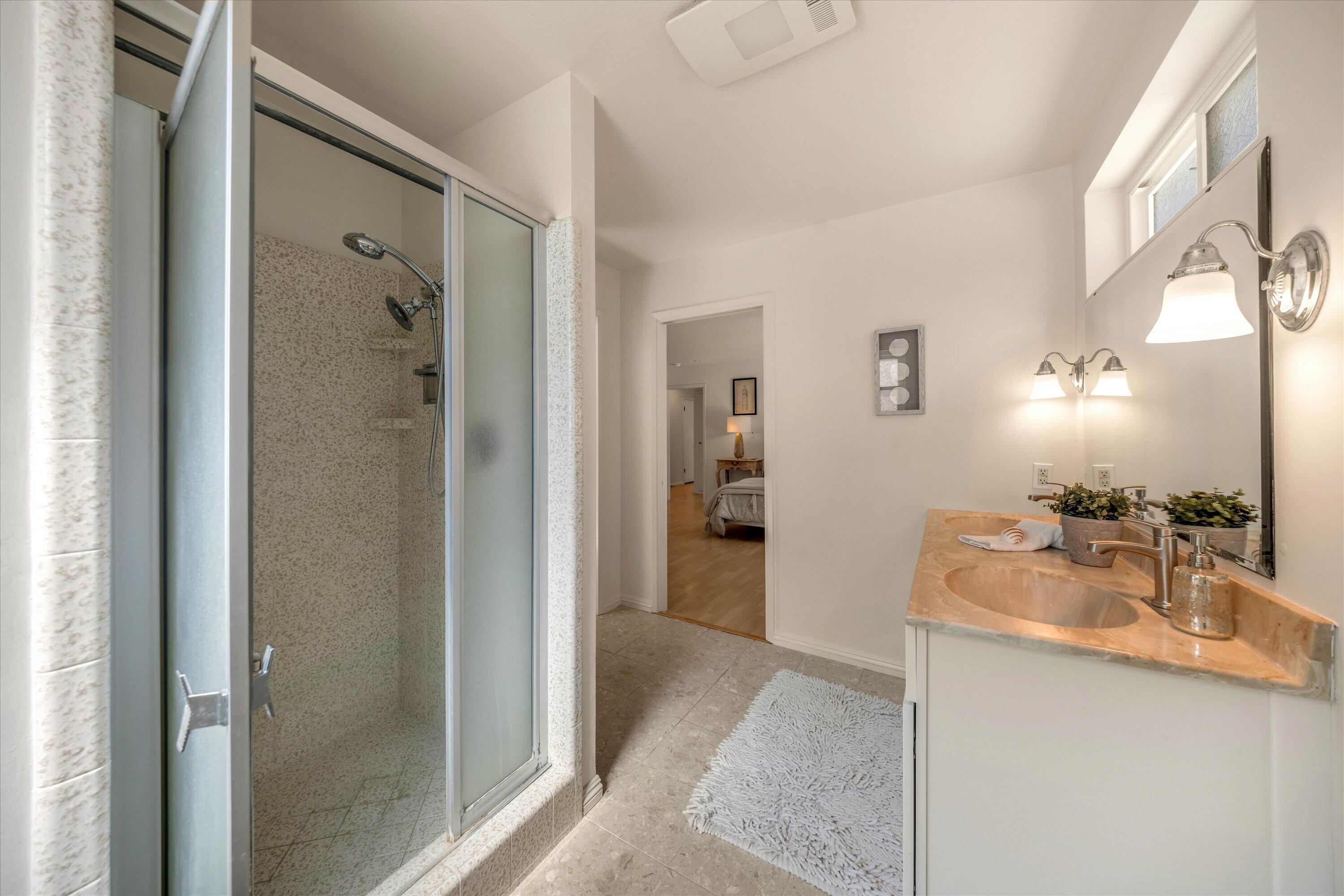 Bathroom with glass shower and double-sink stone vanity beside a doorway to the bedroom.