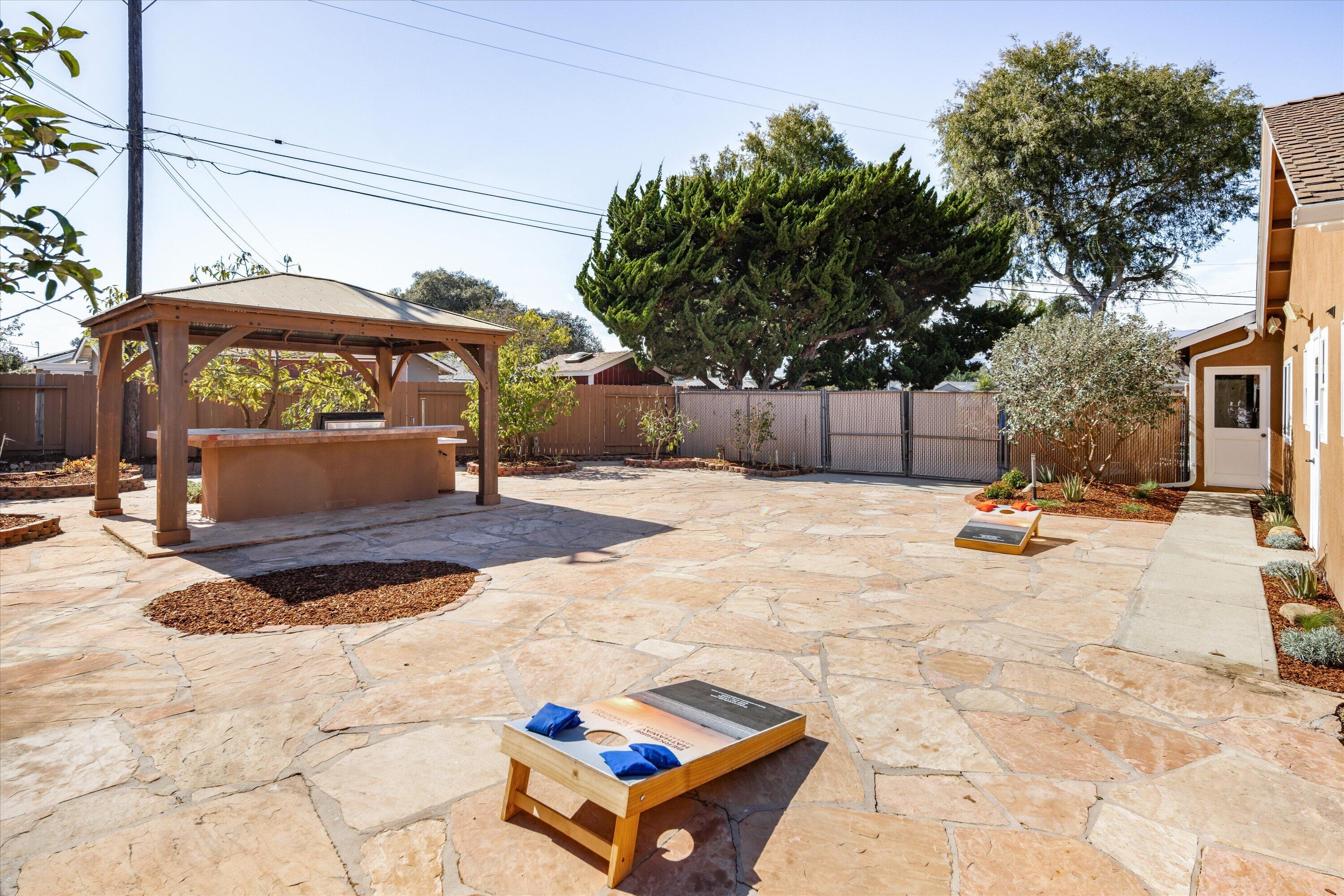 Stone-paved backyard patio with a pergola-covered outdoor kitchen and cornhole game.