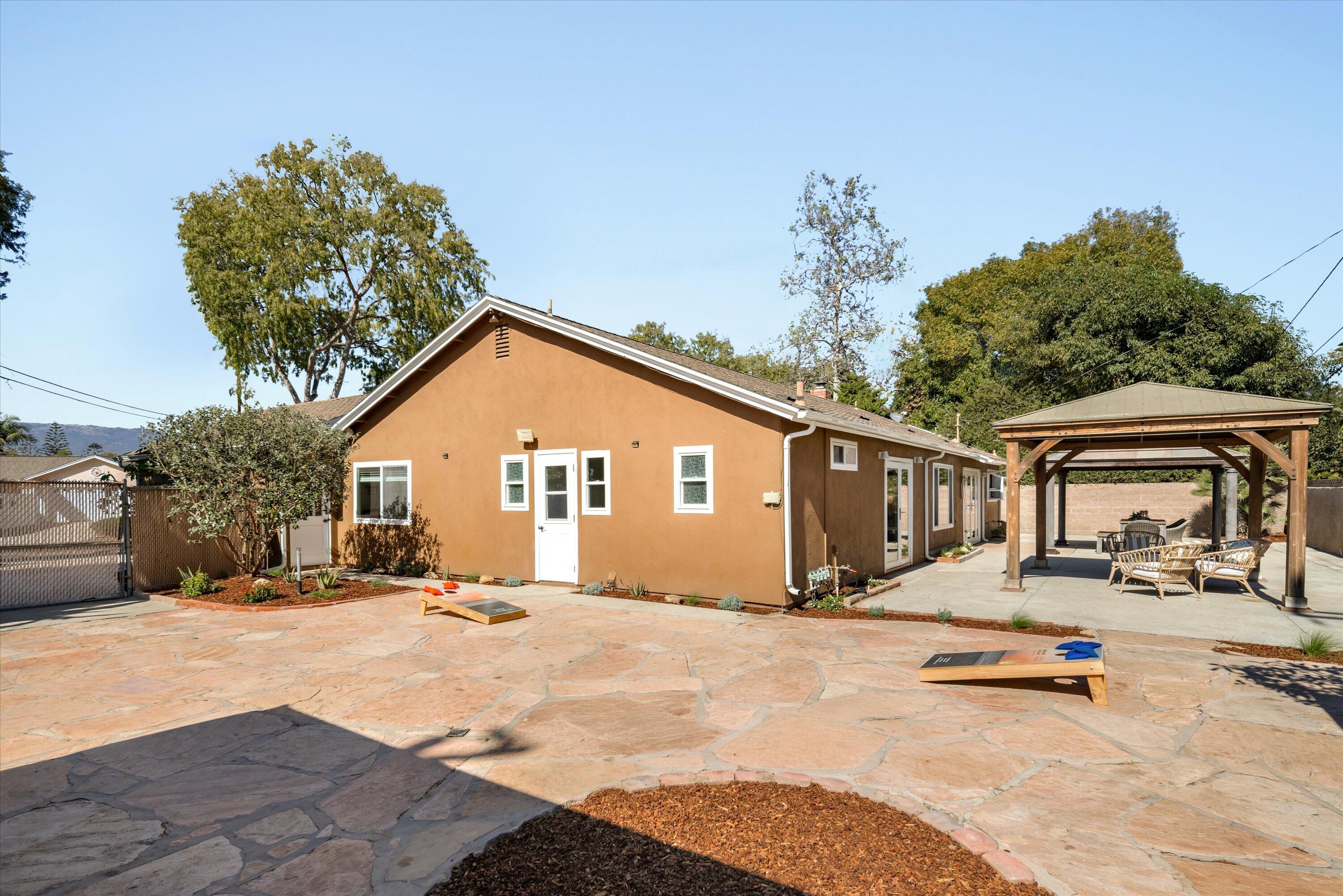 Tan single-story home with a flagstone patio, wooden pergola, outdoor seating, and cornhole boards.