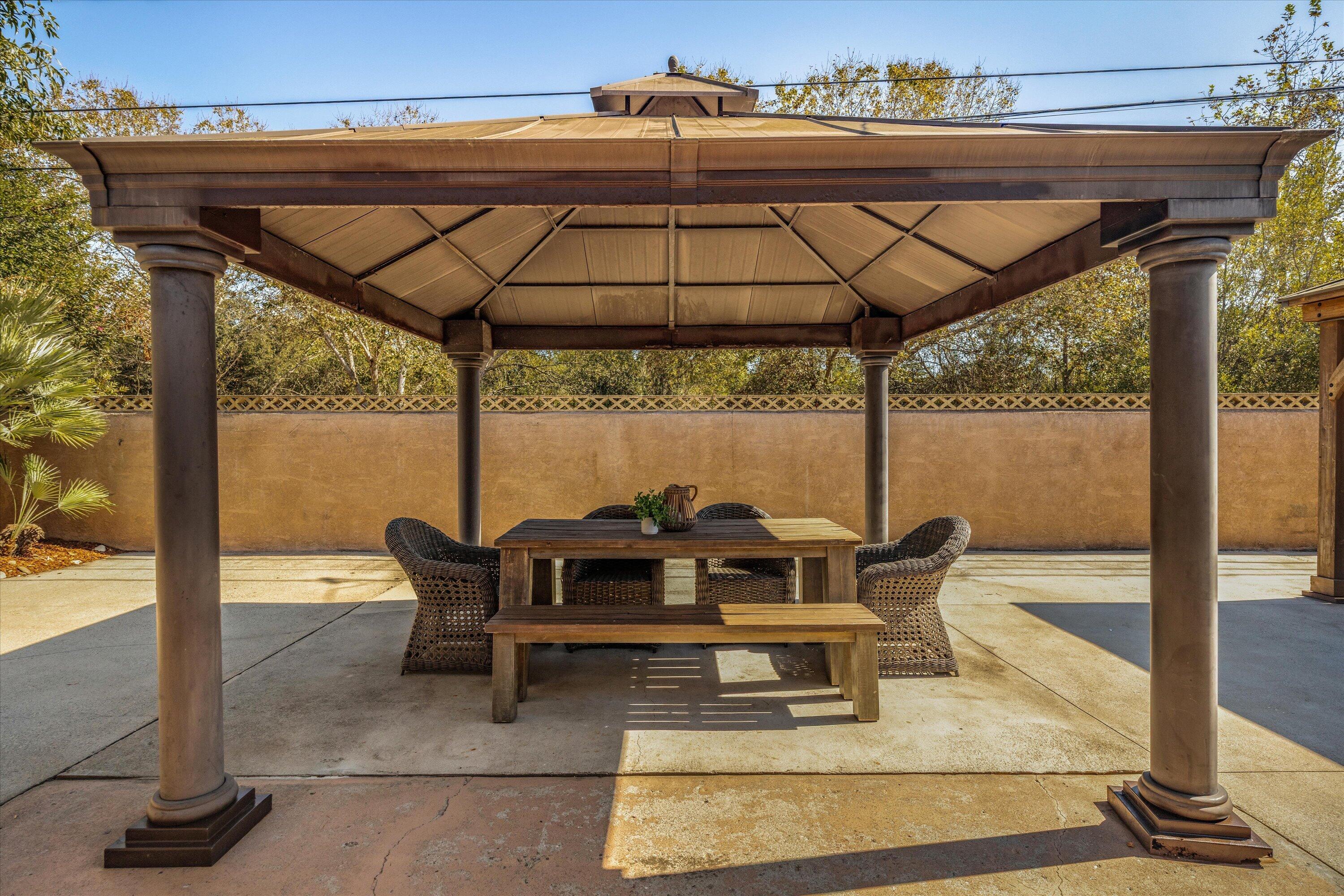 Covered patio with a wooden dining table, bench, and woven chairs beneath a pavilion.