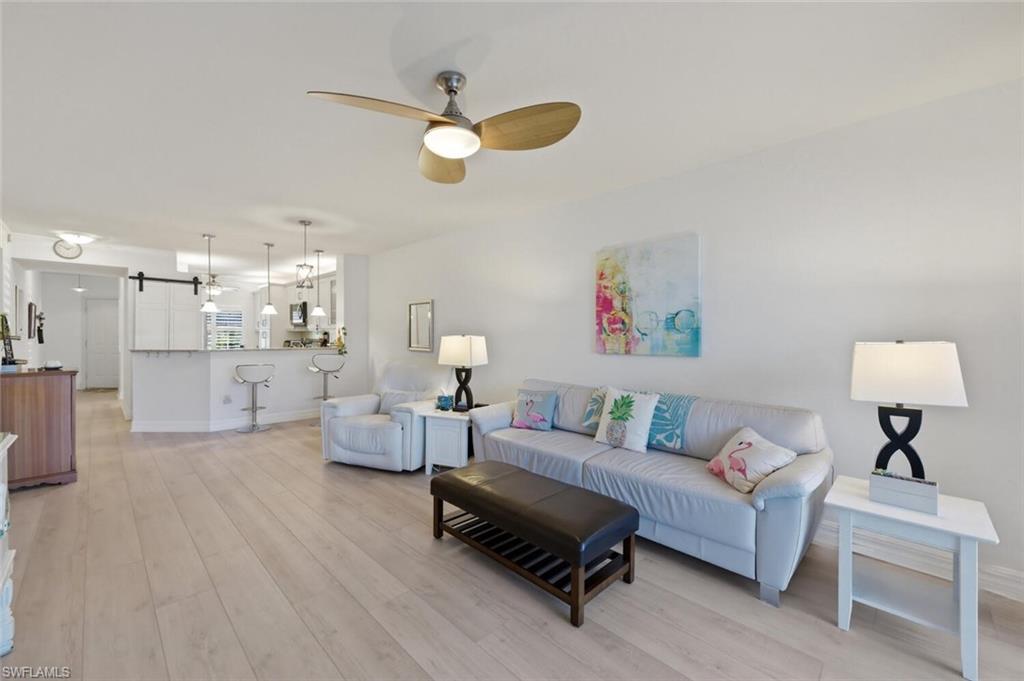 Open-plan living room with a white sectional, pale wood floors, and an adjoining kitchen with breakfast bar.