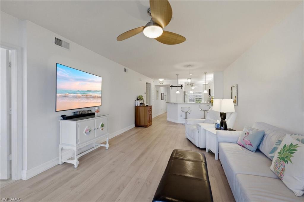 Furnished living room with a white sectional, pale wood floors, and an open kitchen beyond.